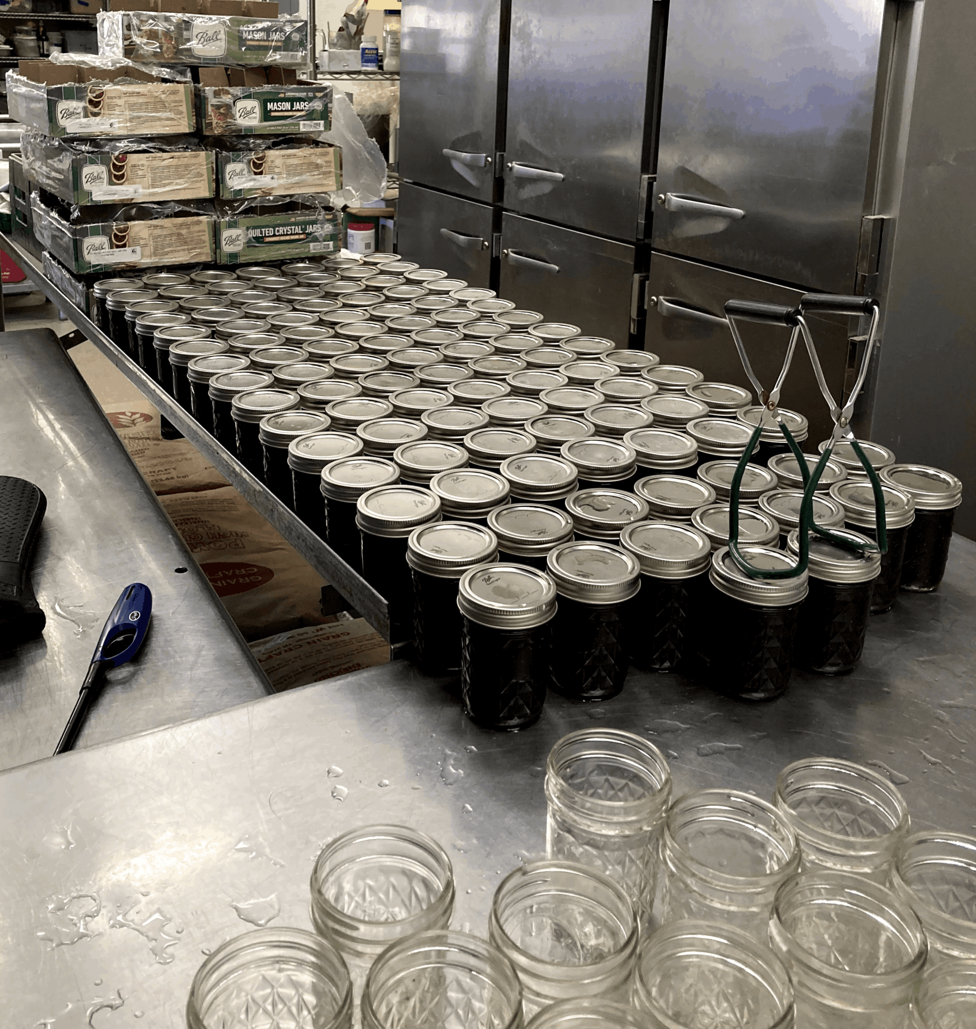 Rows of jars with dark liquid and metal tongs on a stainless steel table in a commercial kitchen.
