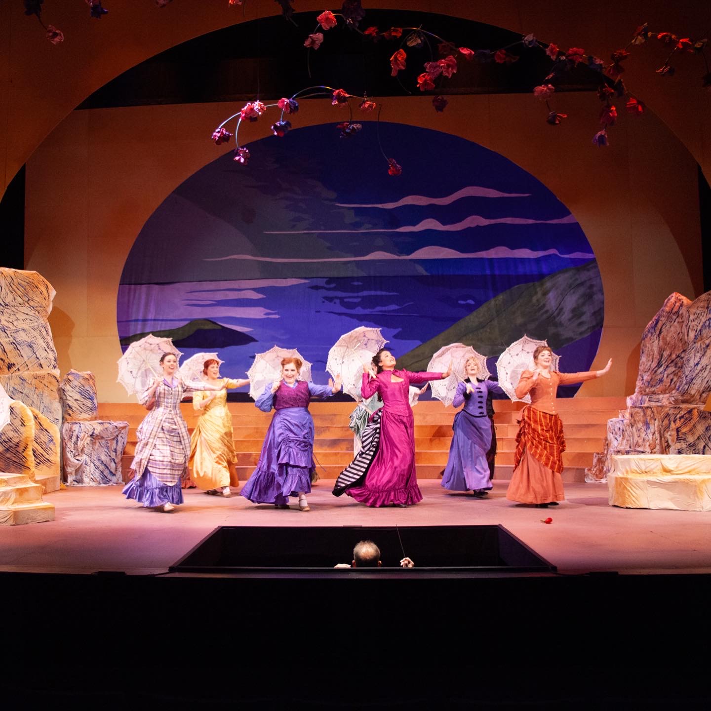 Six performers in colorful costumes dance on stage with a scenic backdrop of mountains and sky, surrounded by rocks and pink flowers.