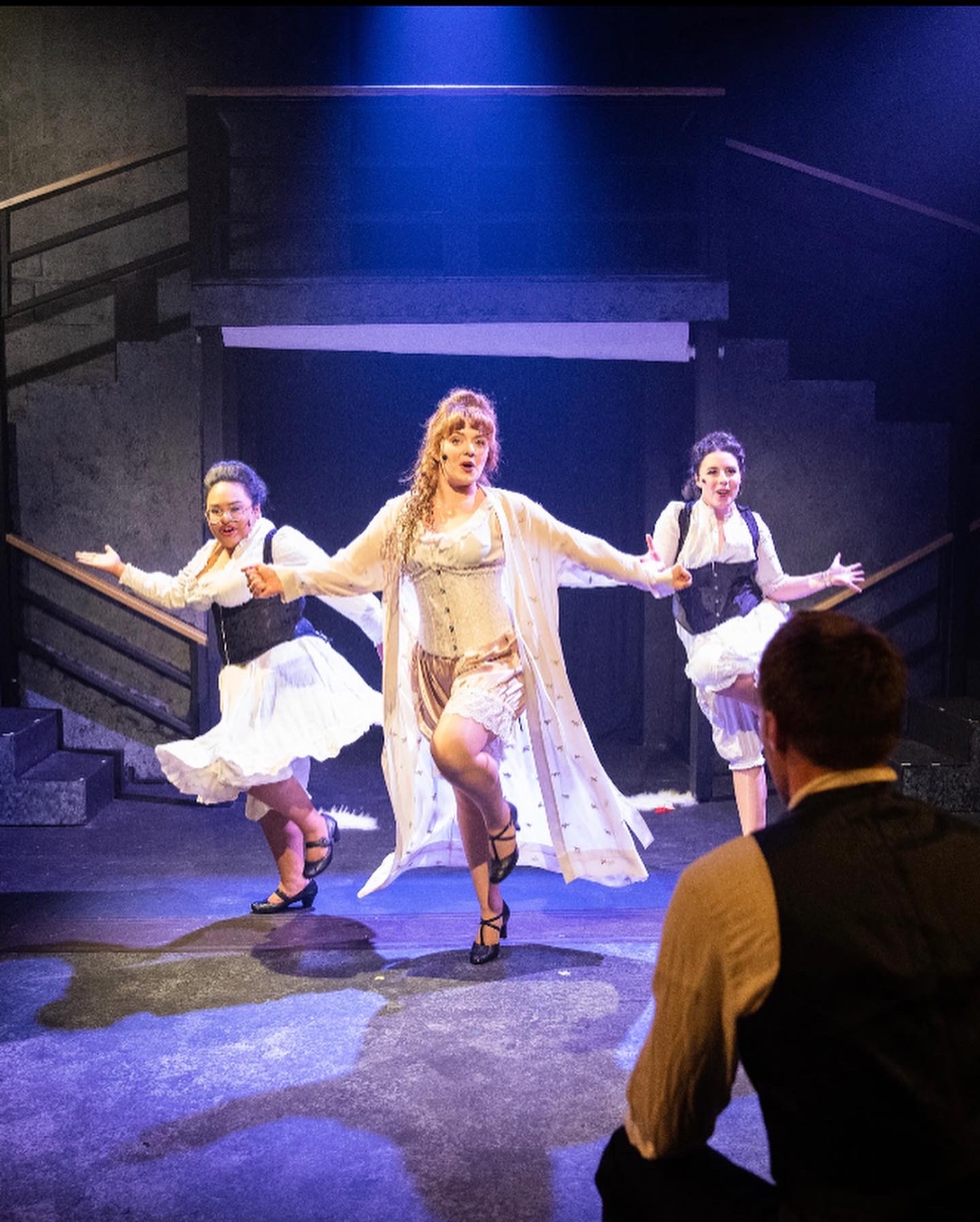 Three women dancing on stage with a man watching, stage set with stairs and dark background, women in white dresses, center woman with arms outstretched.