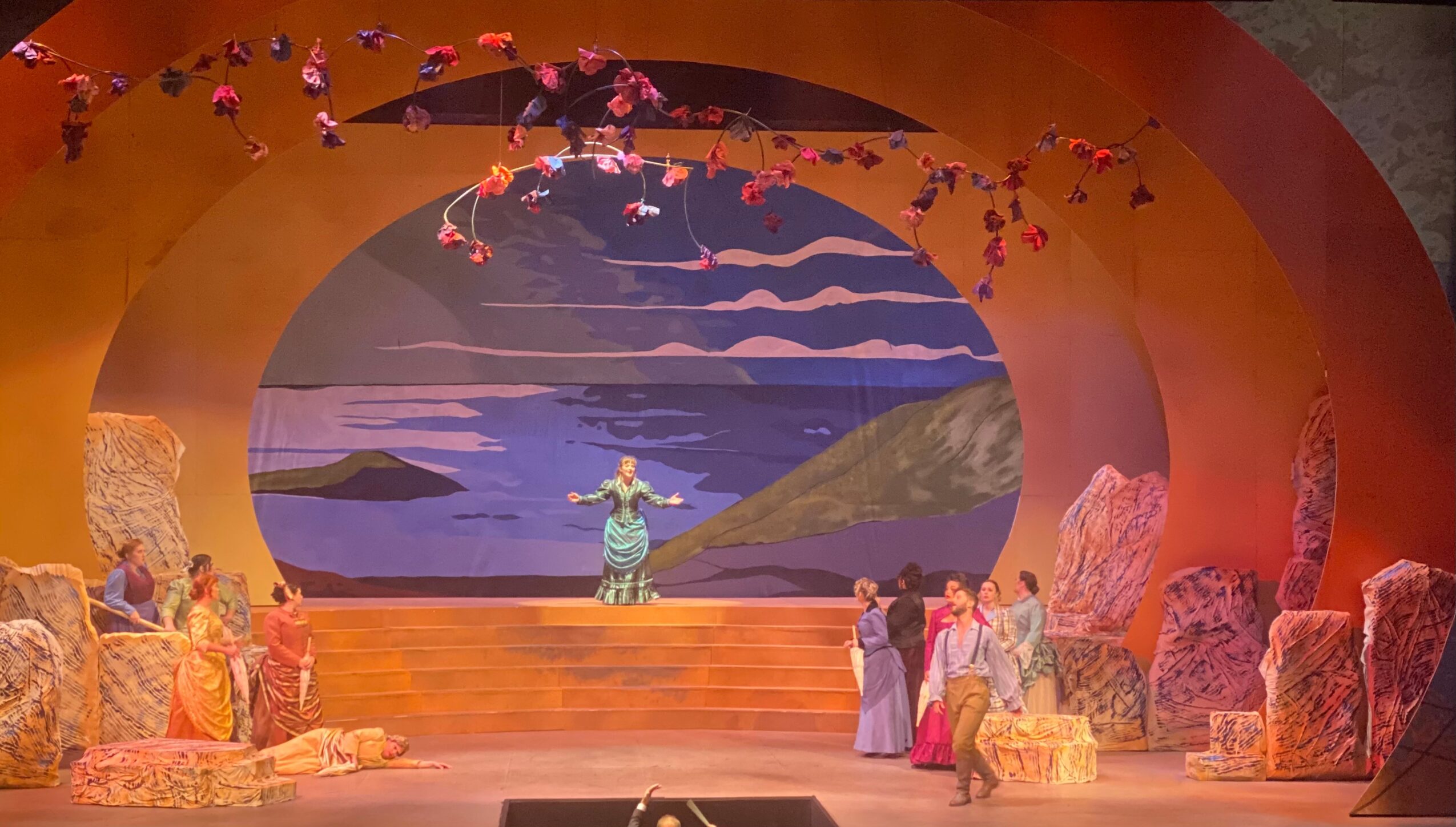 Stage with a central figure in front of a backdrop of water and mountains, surrounded by performers in colorful costumes.