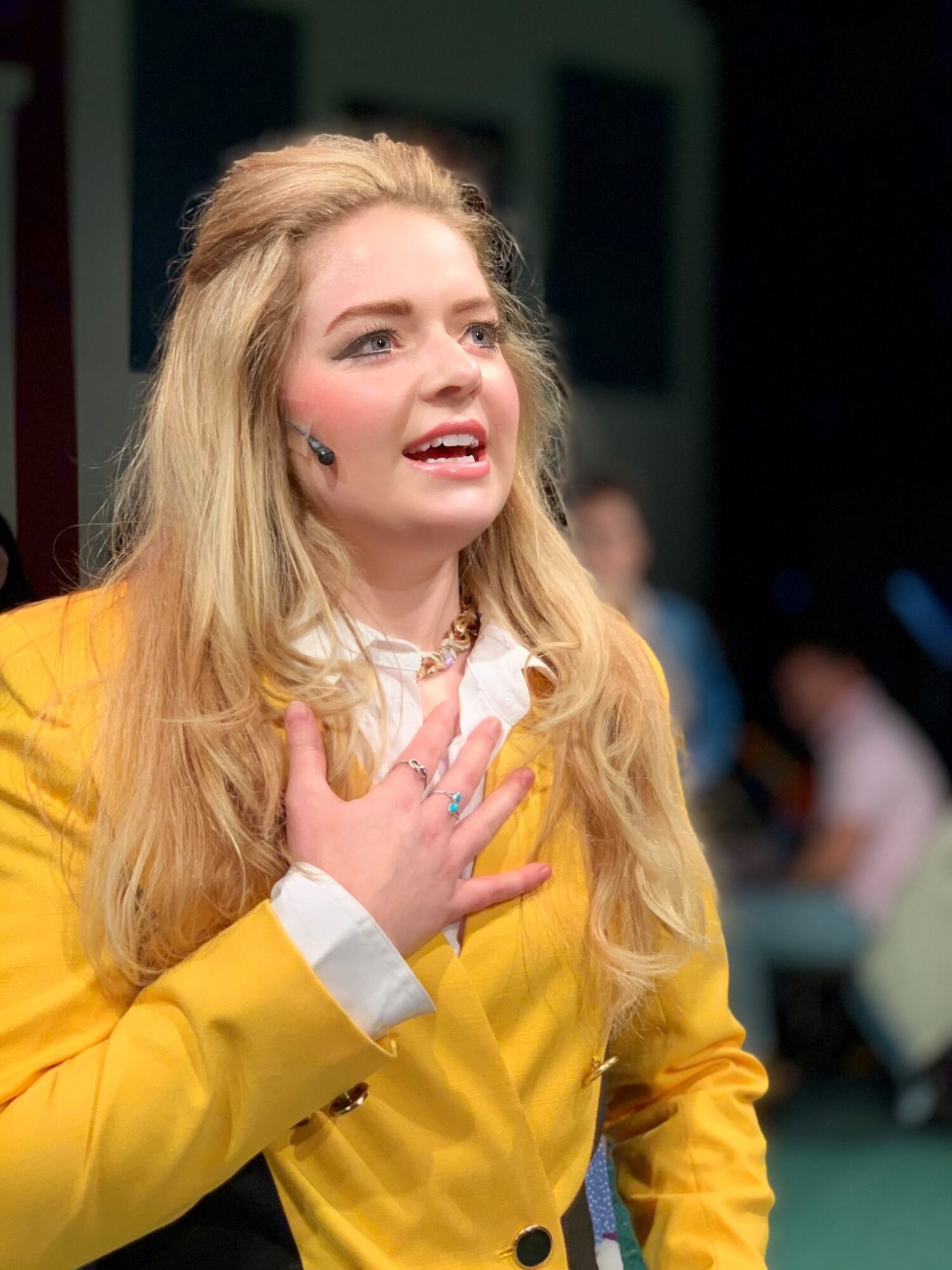 Young woman with long blonde hair wearing a yellow blazer and white shirt, holding her hand to her chest, speaking or singing.
