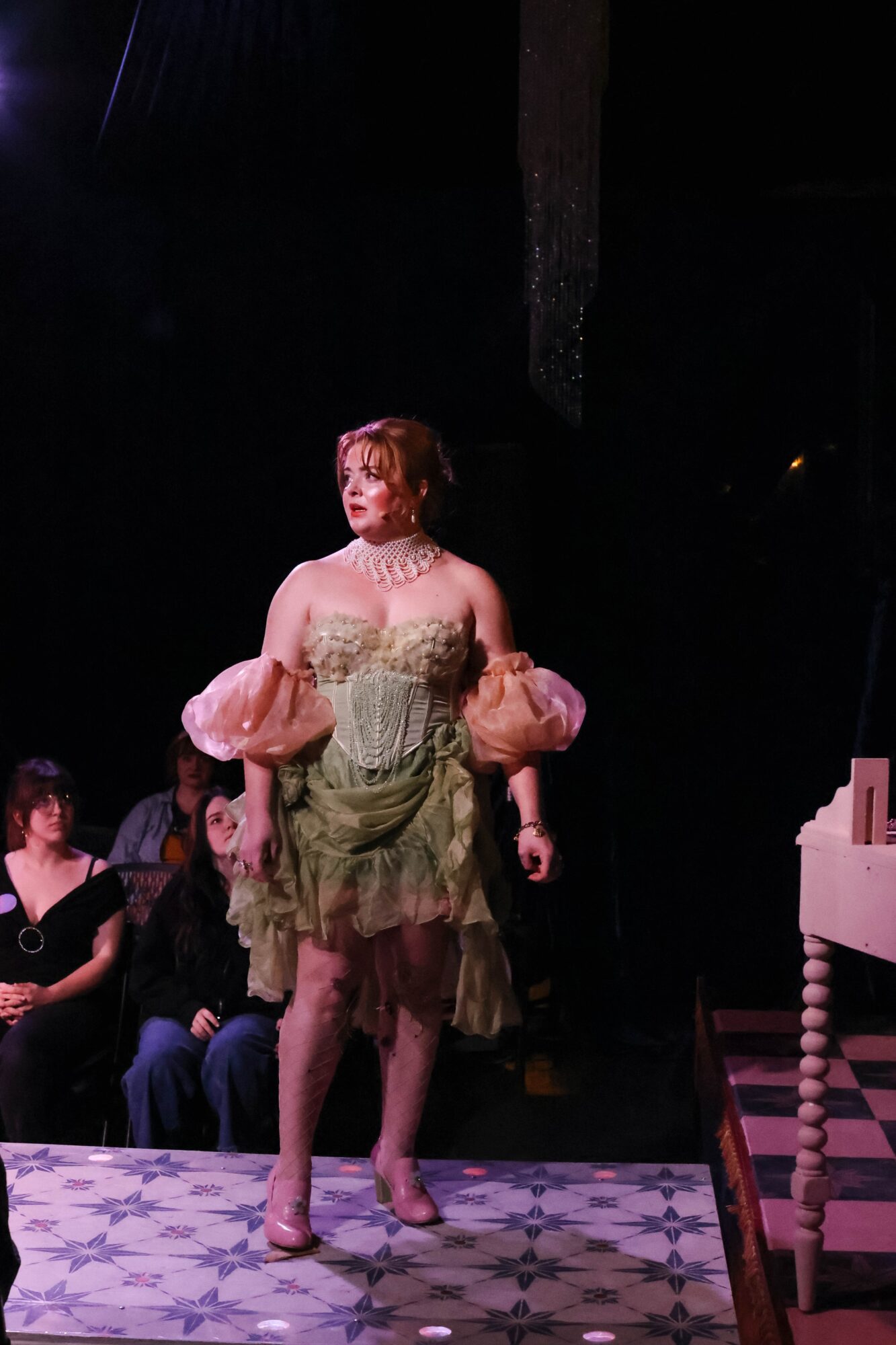 Performer on stage wearing a vintage-style dress with puffy sleeves, standing on a patterned floor, with audience in background.