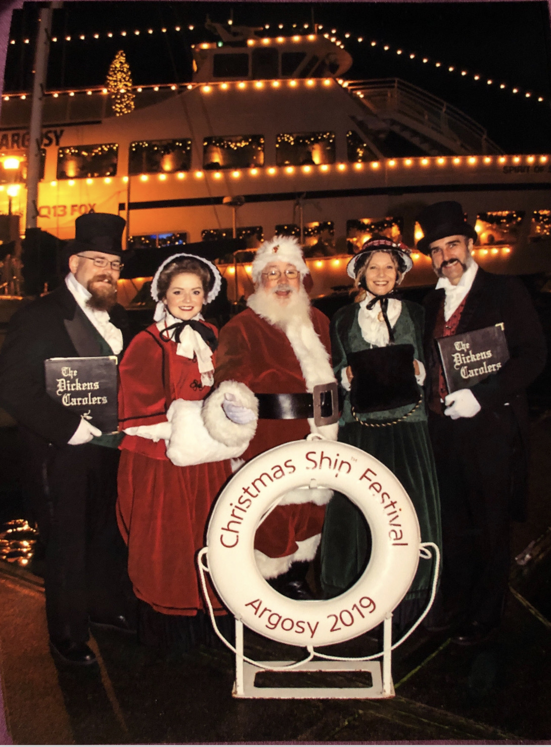 Group of five people dressed as Christmas characters on a boat at night, with festive lights and a sign reading 'Christmas Ship Festival Argosy 2019'.