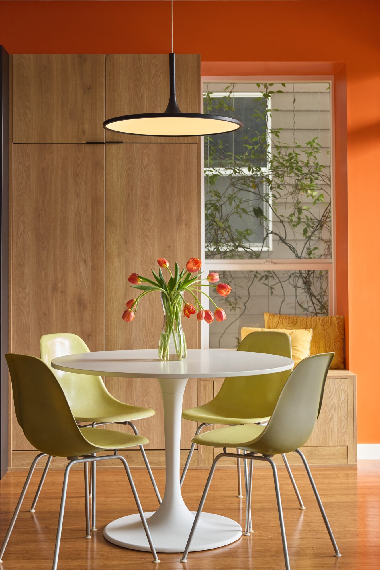 Round dining table with a vase of red tulips, surrounded by six chairs, in a room with wooden cabinets and a window.