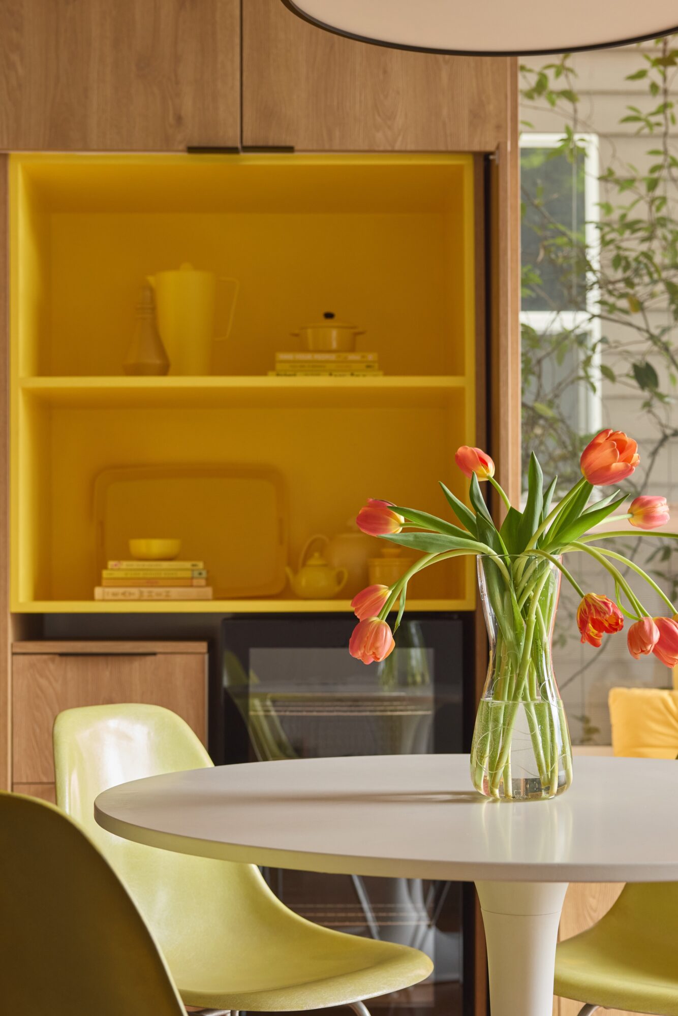 Vase with orange tulips on a white table, yellow cabinet in background, part of a kitchen or dining area.