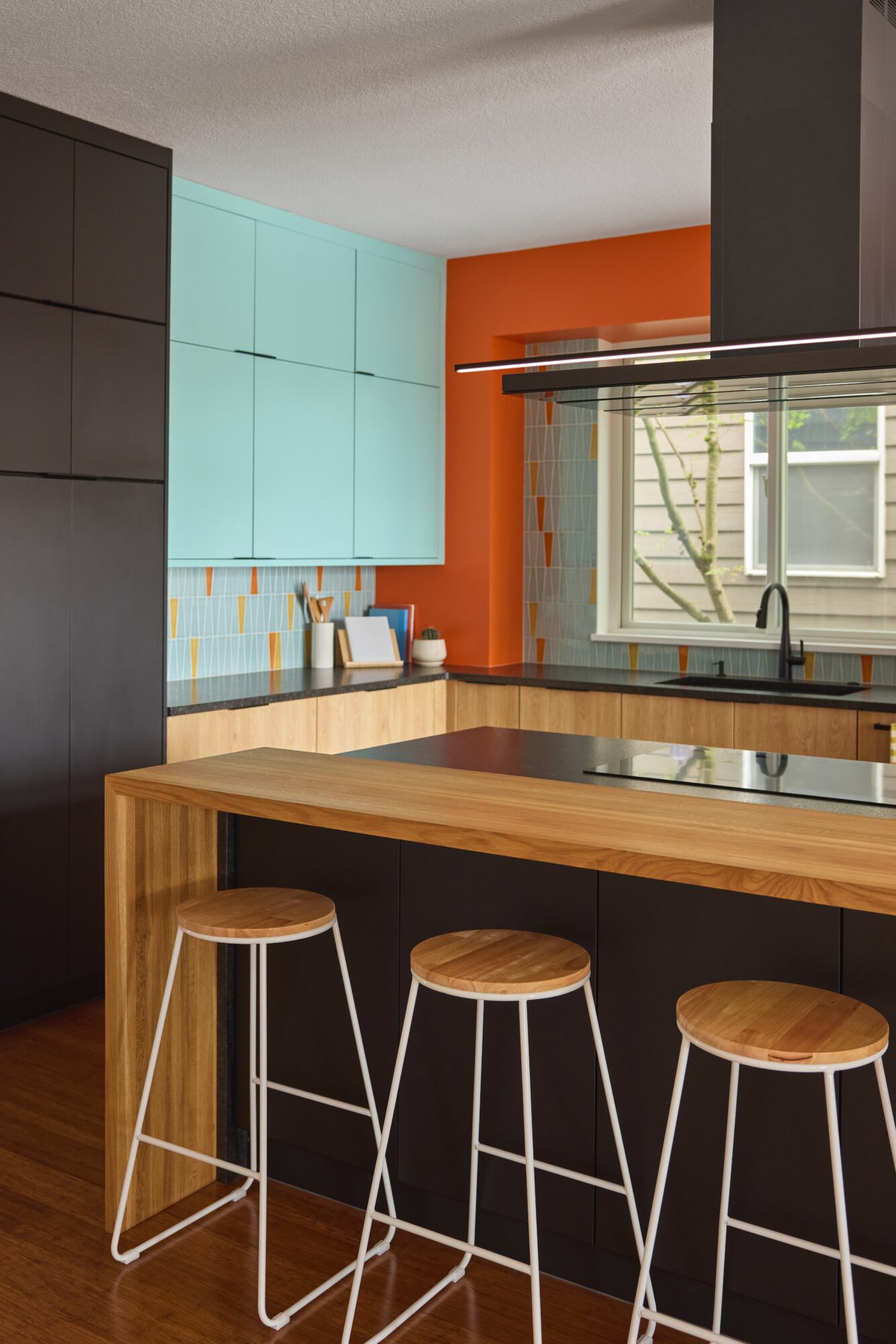 Kitchen with wooden island, three stools, colorful cabinets, window with blinds, and orange wall.