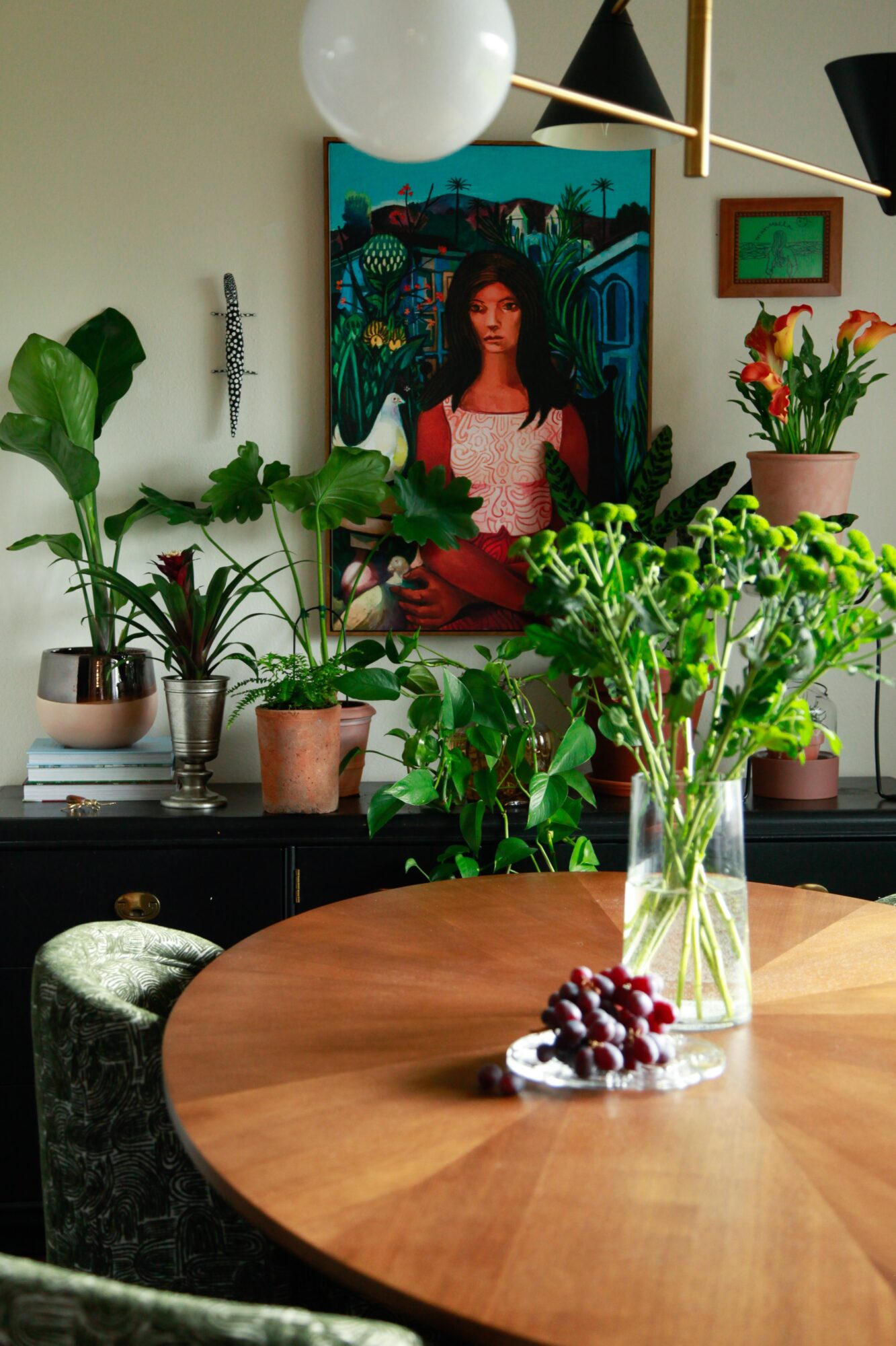 Interior room with plants, a painting of a woman, and a wooden table with a vase of flowers and berries.