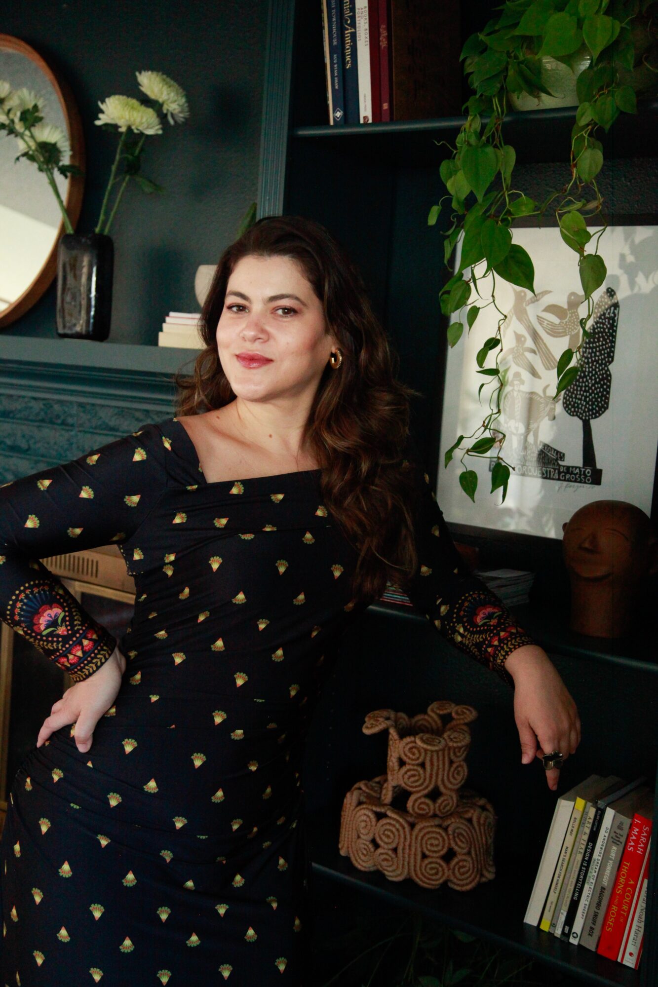 Woman with long dark hair wearing a black dress with gold patterns, sitting in a room with books and plants.
