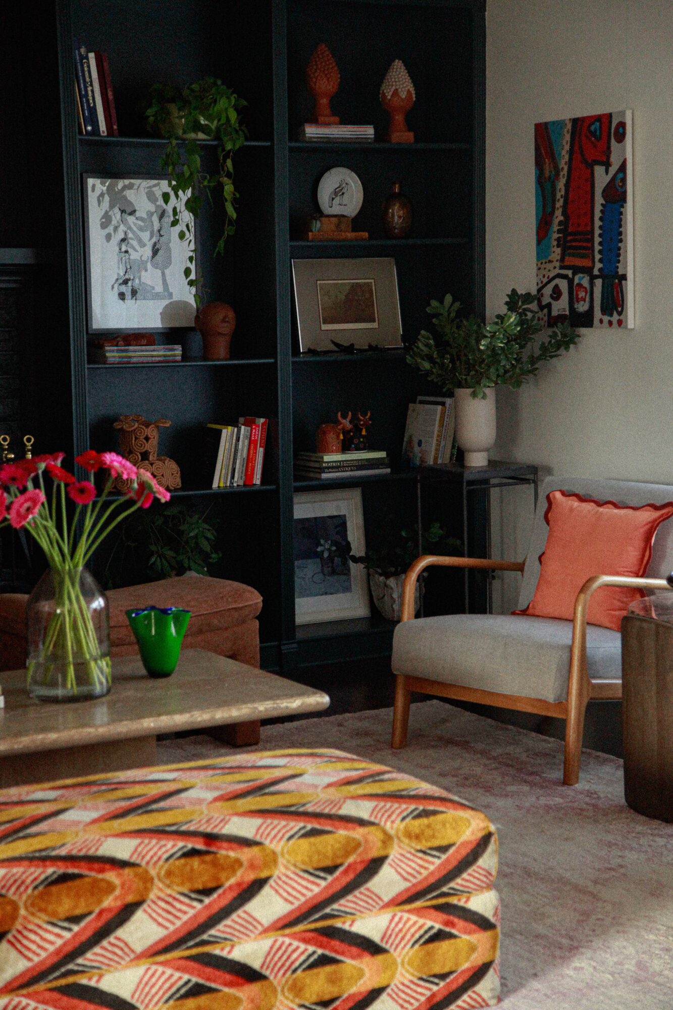 Living room with a dark bookshelf, armchair with pink cushion, wooden coffee table, and colorful wall art.