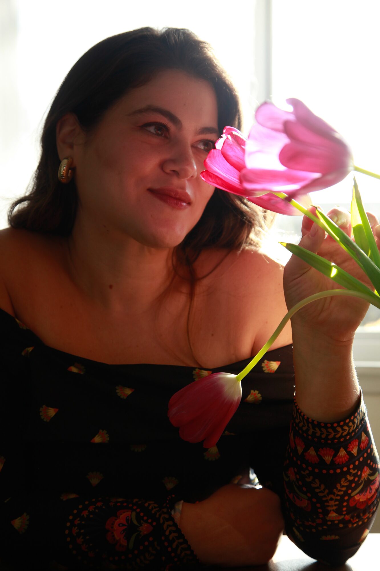 Woman with dark hair holding pink and red tulips, smiling, indoors near a window, wearing a black top with gold accents.