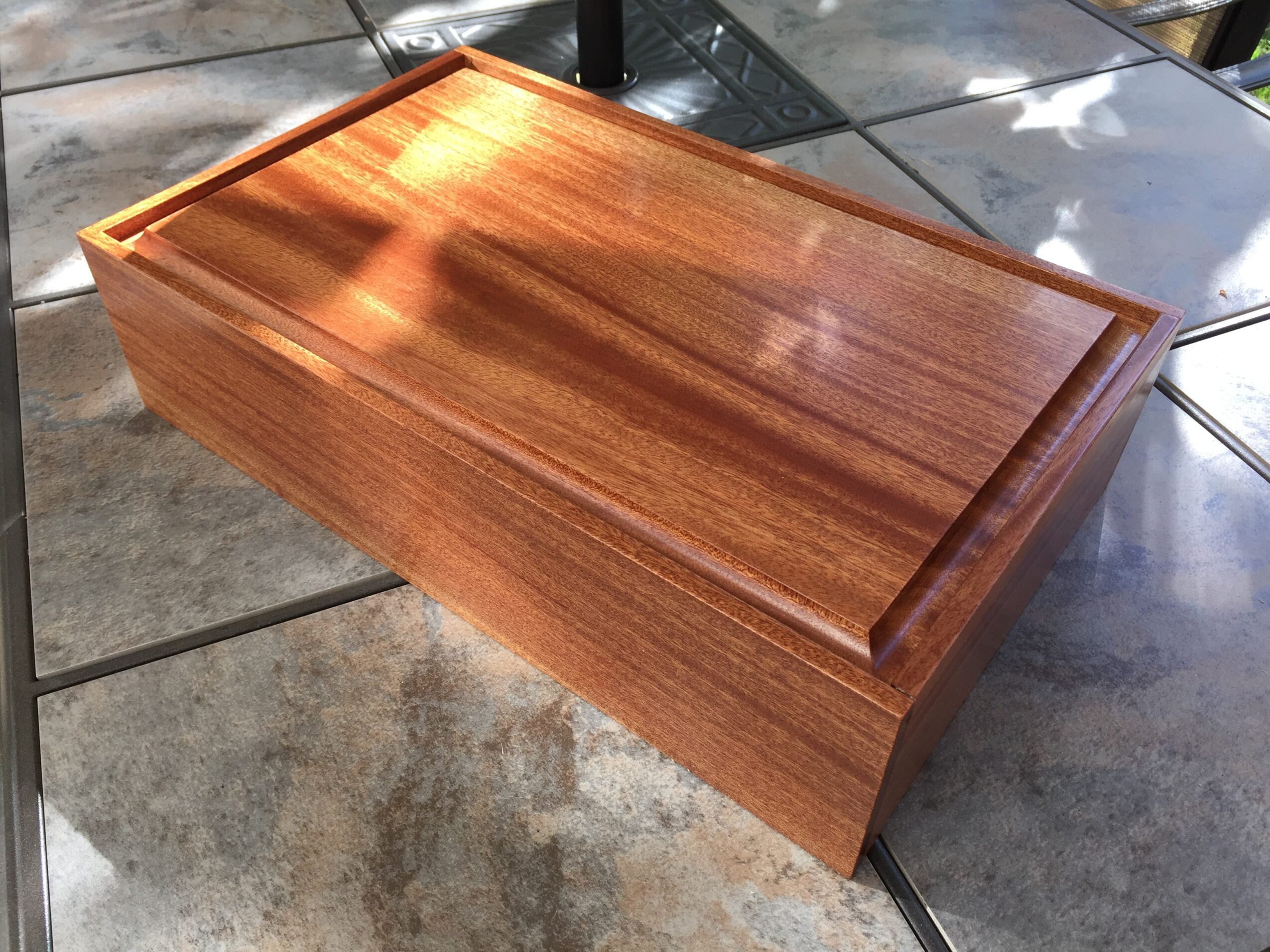 Rectangular wooden box with a smooth, polished surface and a raised border on the top, placed on tiled floor.