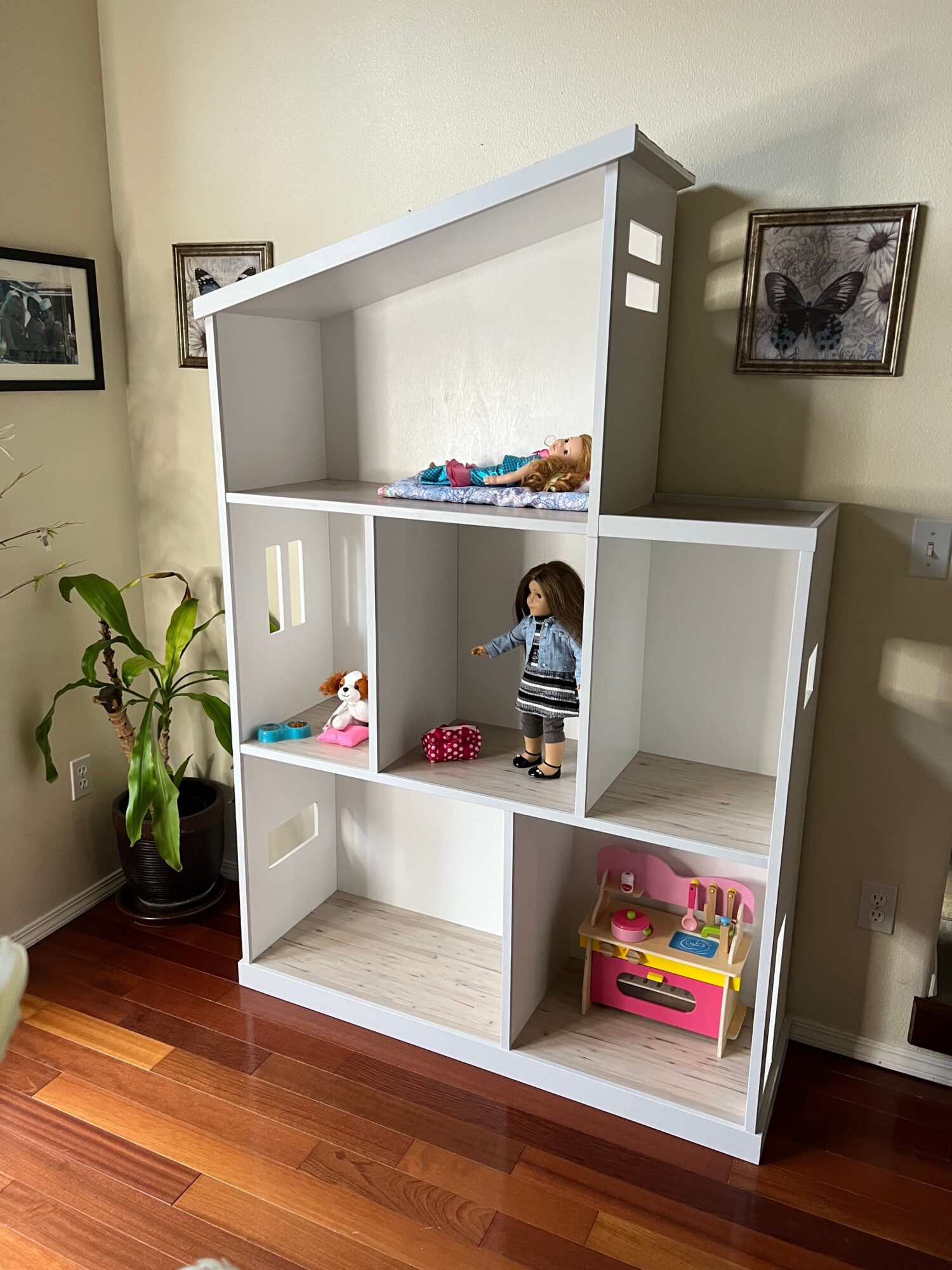 Child standing in a white bookshelf with colorful toys and books, next to a potted plant and framed pictures on the wall.