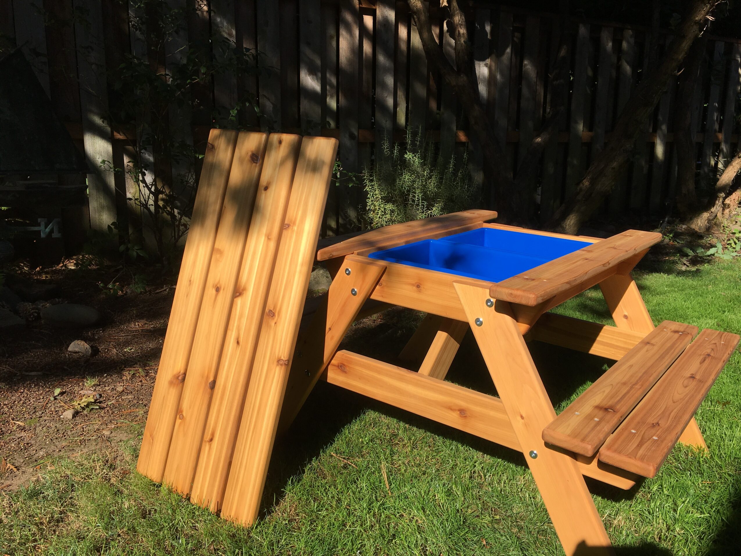 A wooden sandbox with a blue interior, a wooden lid leaning against it, and a small wooden bench on grass.