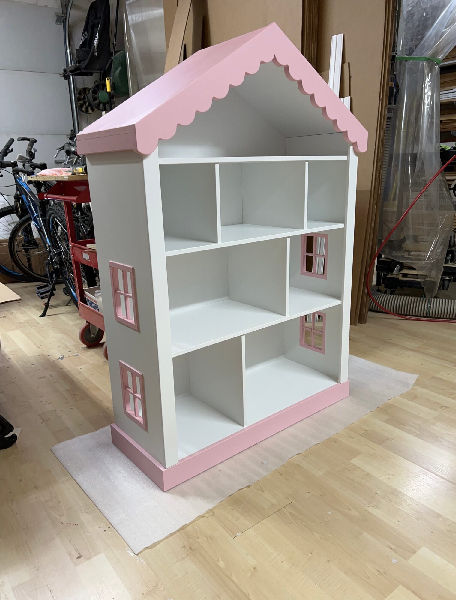 Pink and white dollhouse shelf with windows and a scalloped roof, placed on a wooden floor.
