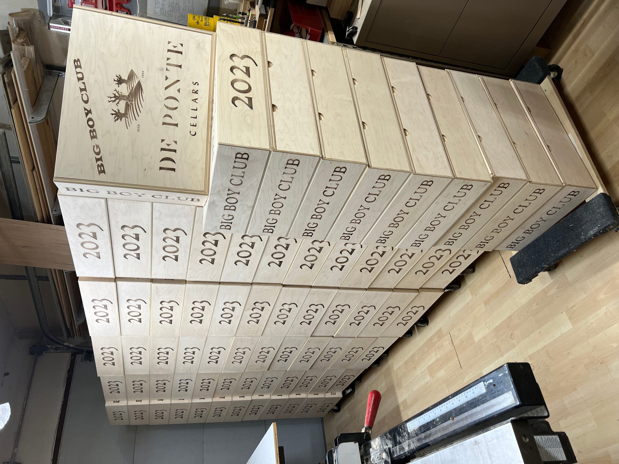 Stack of white boxes with black text and logos, arranged vertically on a wooden floor, with a black object and a red handle nearby.