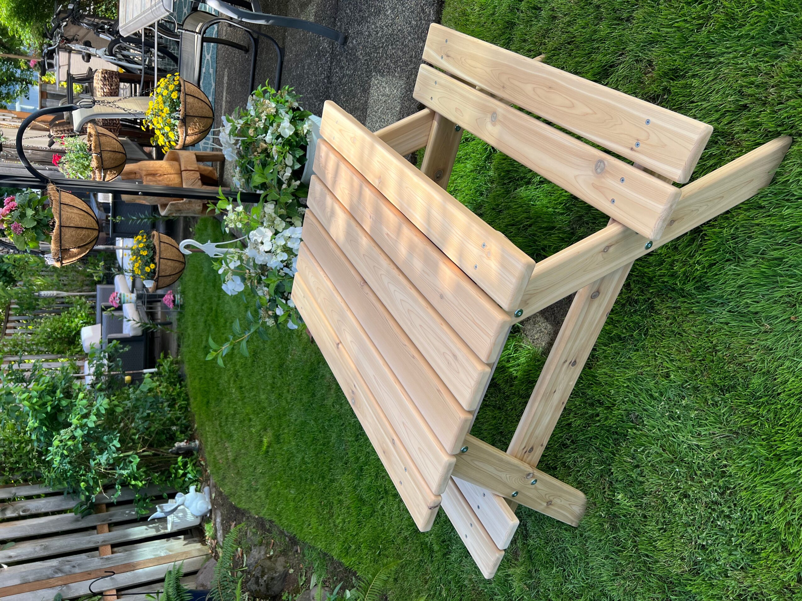 Wooden outdoor table with attached bench on a grassy lawn, garden and outdoor furniture in background.