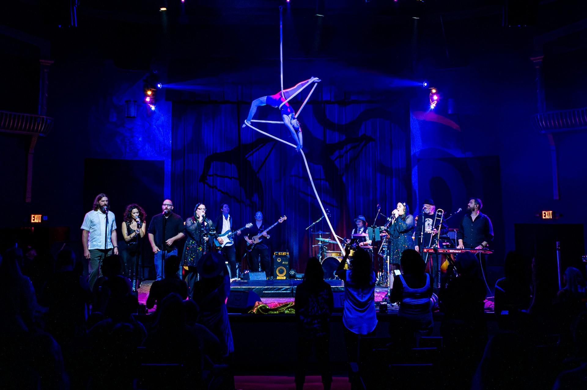 Band performs on stage with audience watching, stage lit with blue and purple lights, large geometric sculpture above.
