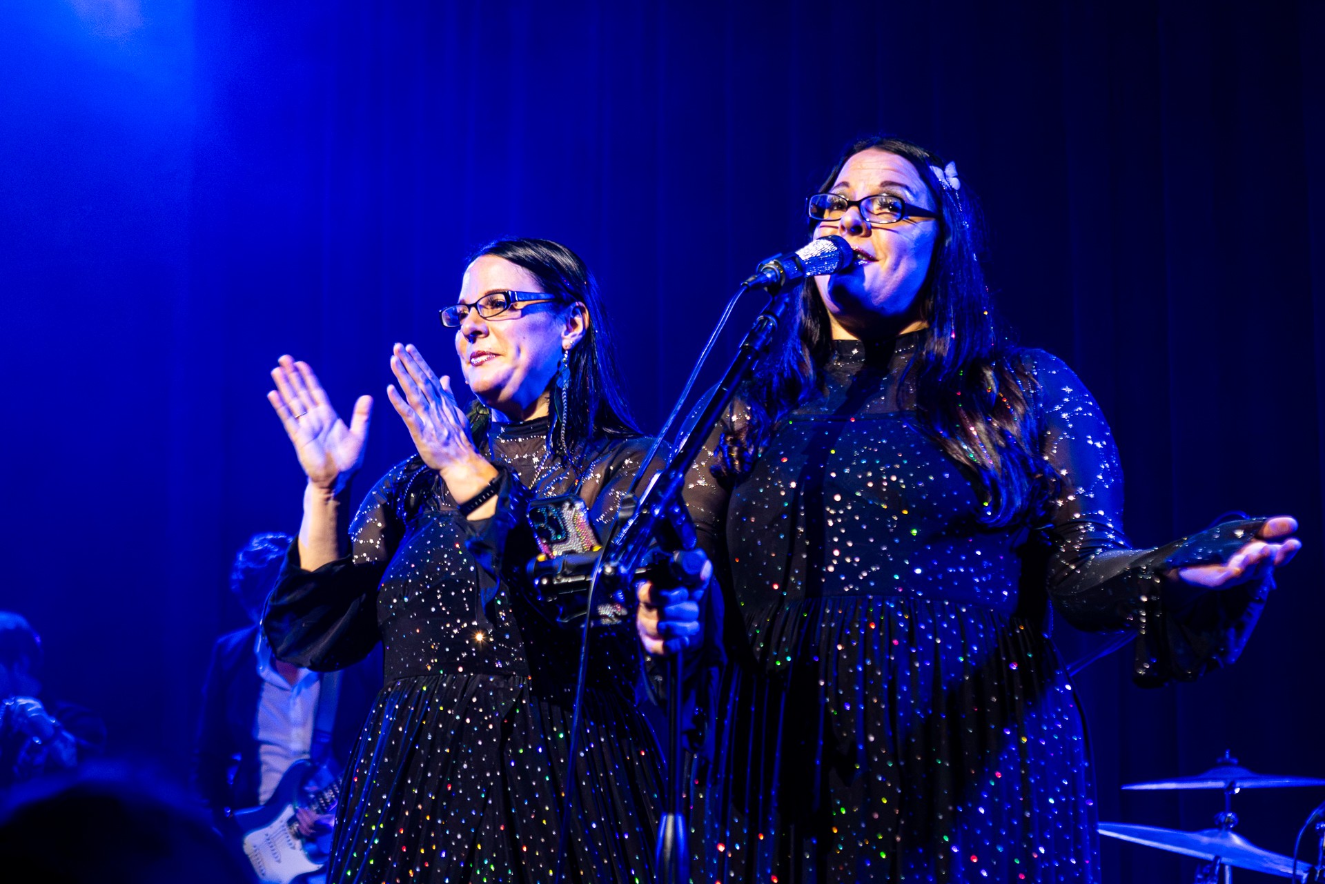 Two women singing on stage with microphones, wearing matching sparkly outfits, under blue stage lighting.