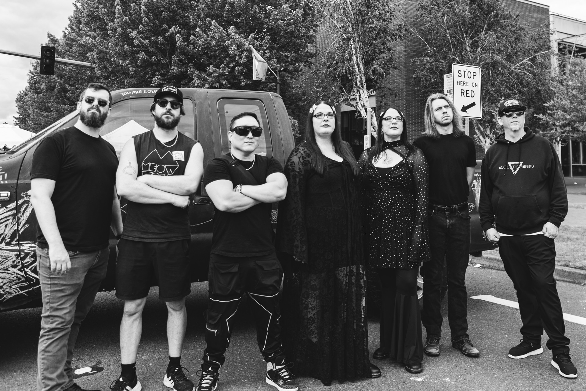 Group of seven people standing outdoors on a street, some wearing sunglasses, with trees and a stop sign in the background.