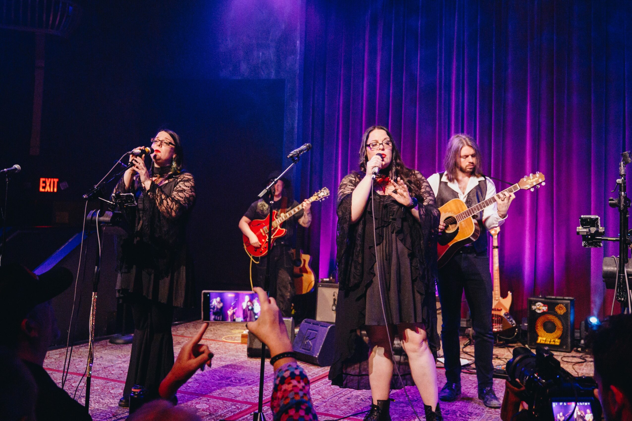 Four women perform on stage with purple lighting, two singing into microphones, one playing guitar, and one with a ukulele, audience taking photos.