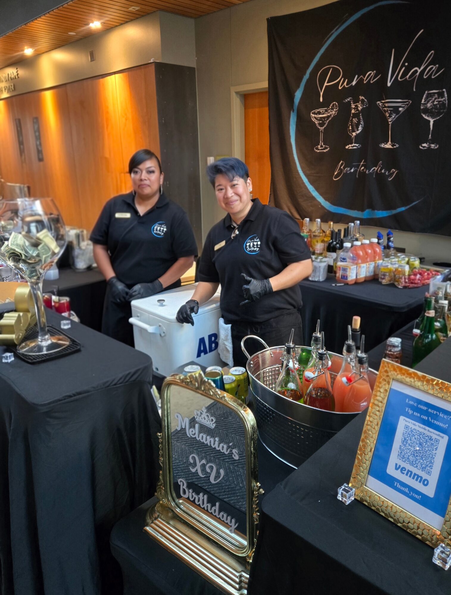 Two women in black uniforms behind a bar setup with drinks and decorations, a banner with cocktail images and text in the background.