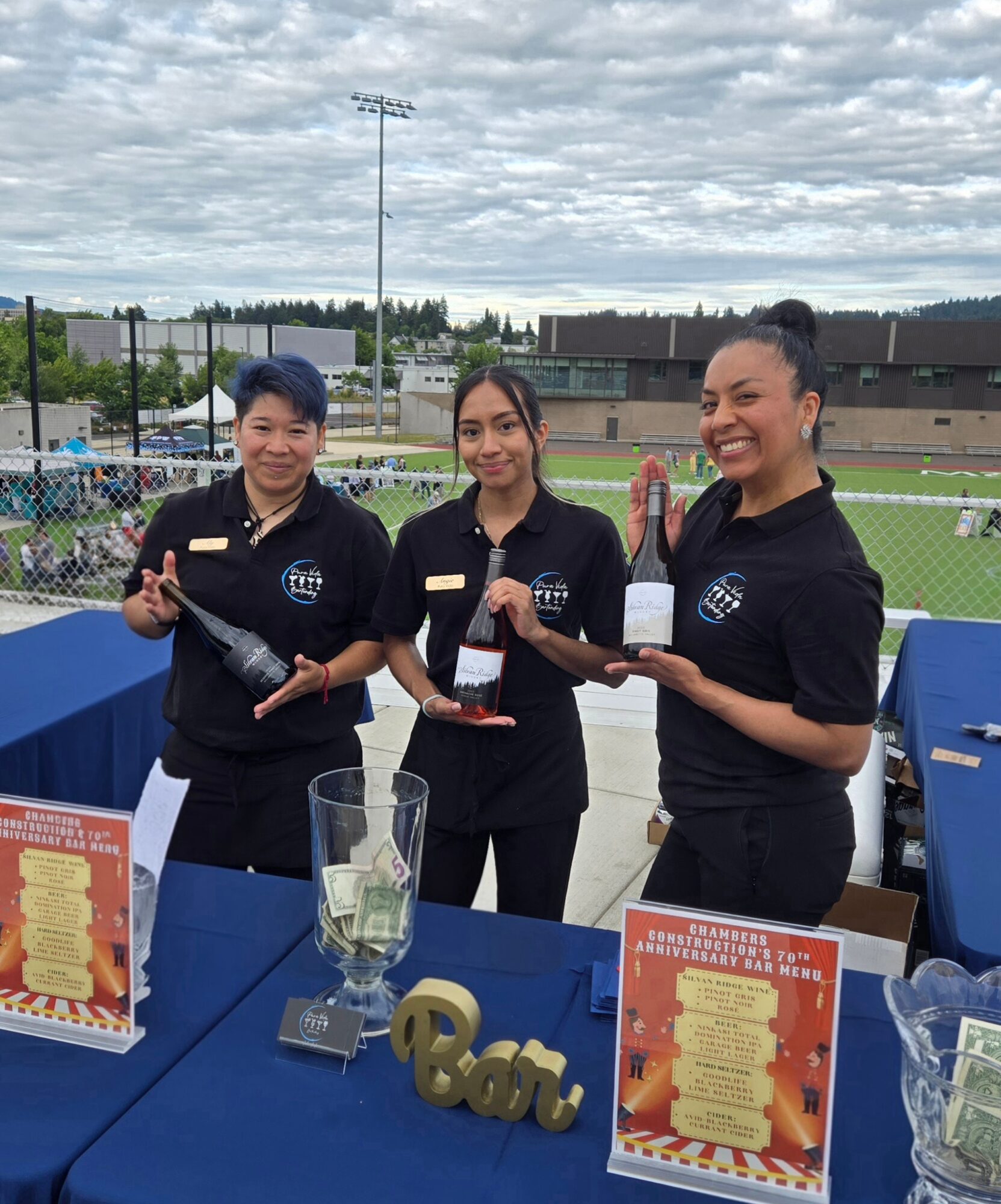 Three women in black uniforms standing behind a table outdoors, holding bottles, smiling, with a sports field in the background.
