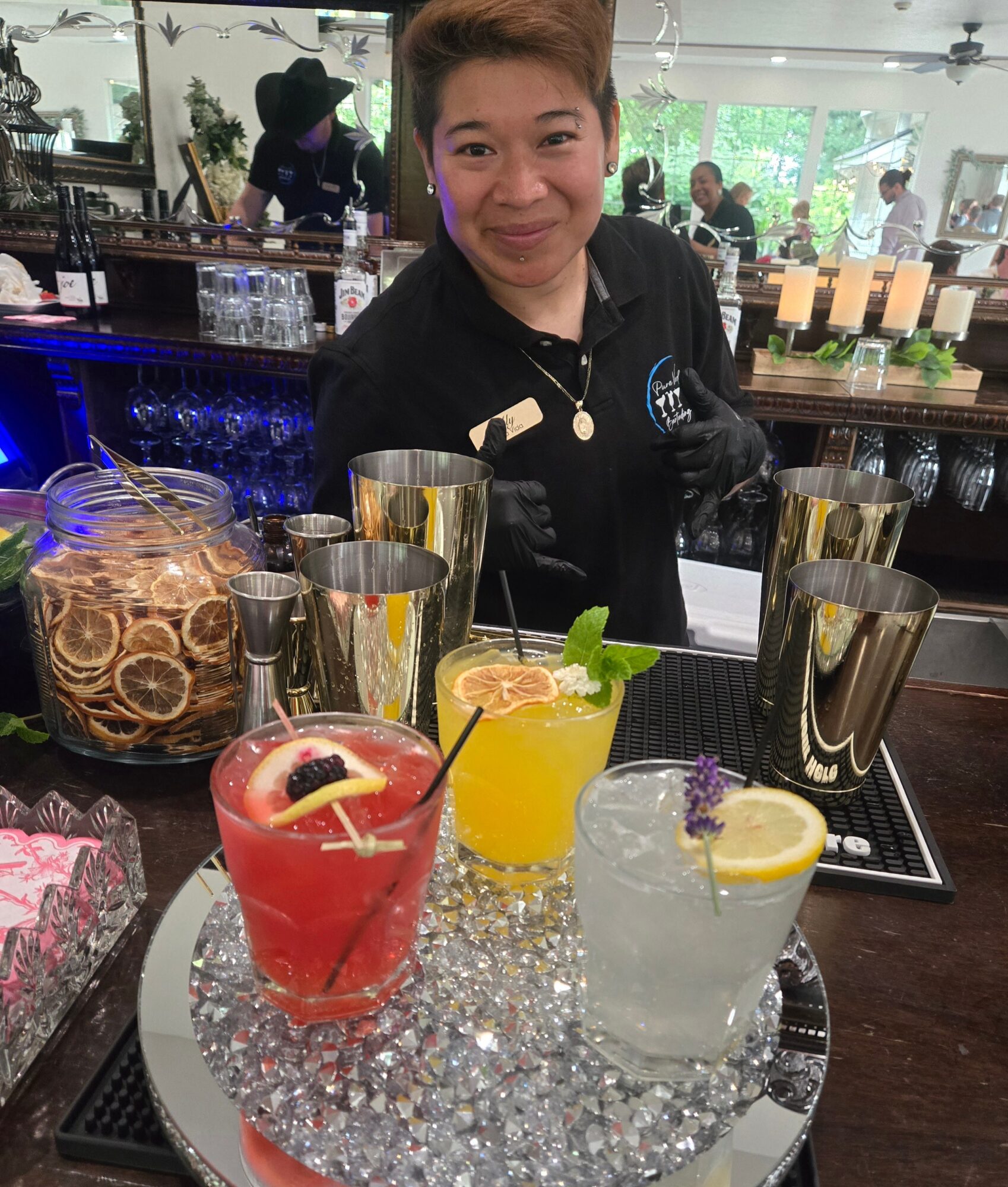 A smiling bartender behind a bar with three colorful drinks on a tray, in a well-lit bar or restaurant setting.