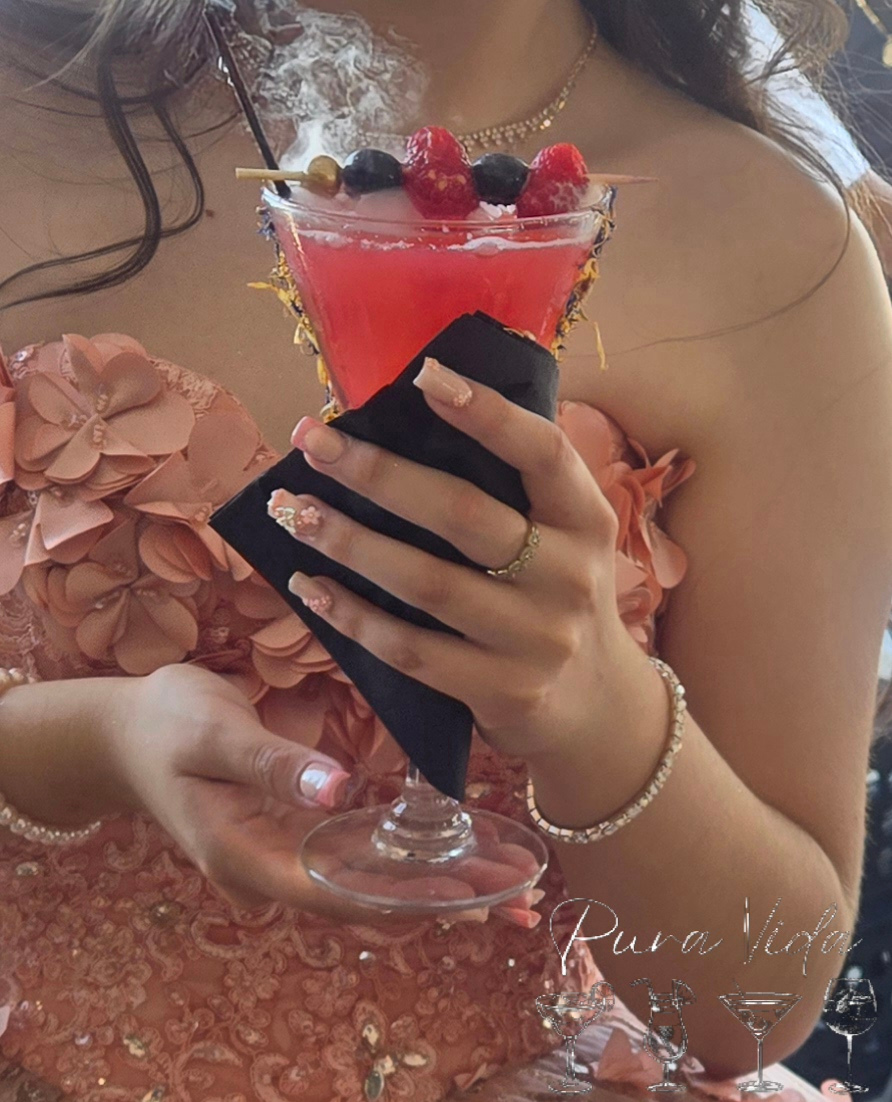 Person holding a pink cocktail garnished with berries, wearing a peach-colored dress with ruffled details, and jewelry.