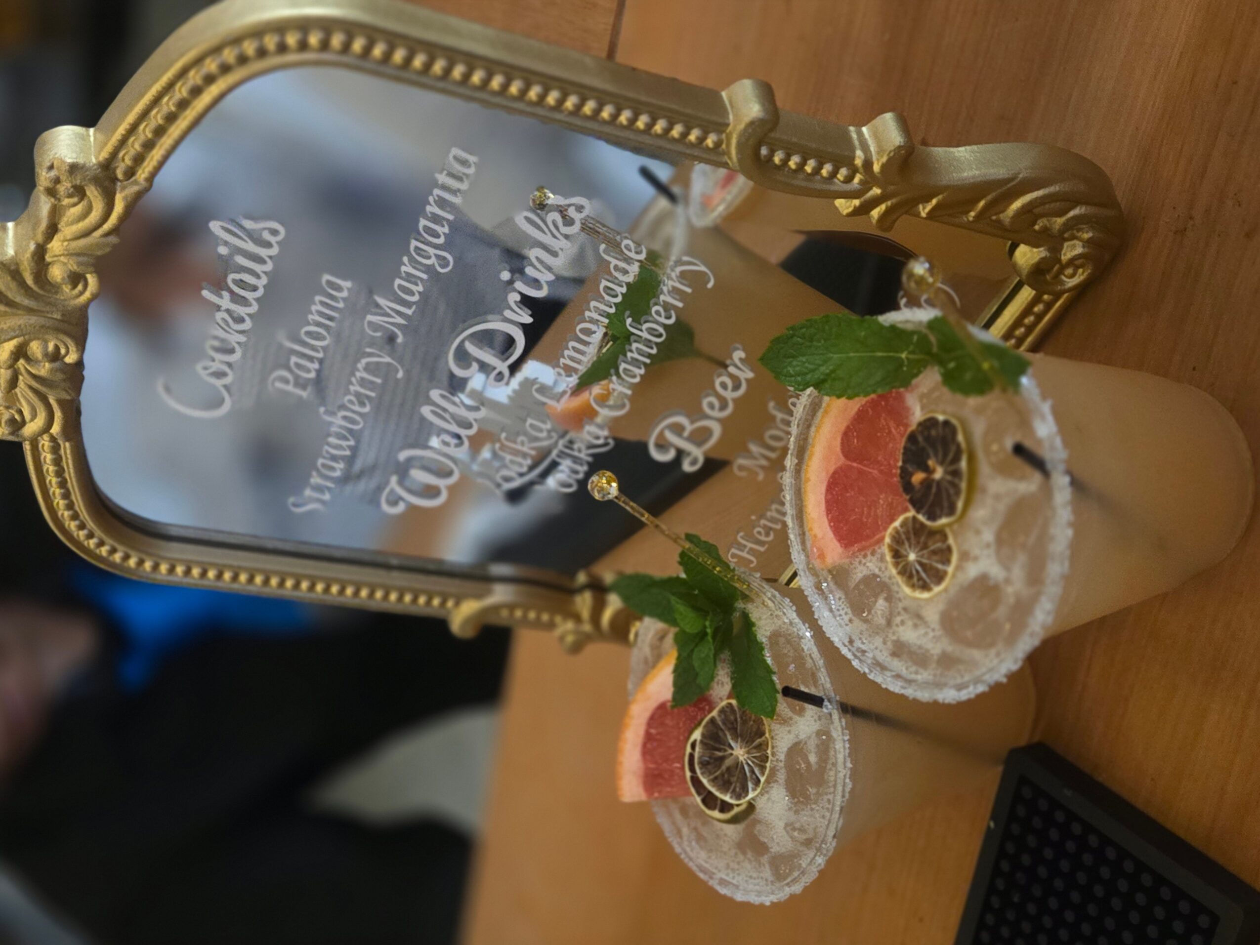 Two cocktails with mint leaves, grapefruit slices, and blackberries on a wooden table, reflected in a mirror with gold frame.