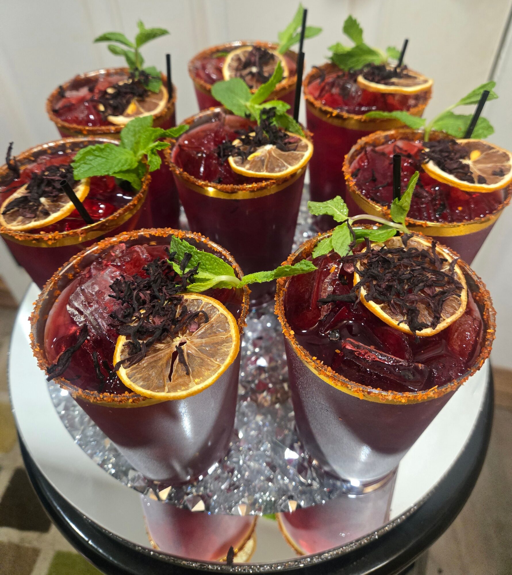 Eight glasses of dark red beverage garnished with lemon slices, mint leaves, and black straws, arranged on a silver tray.