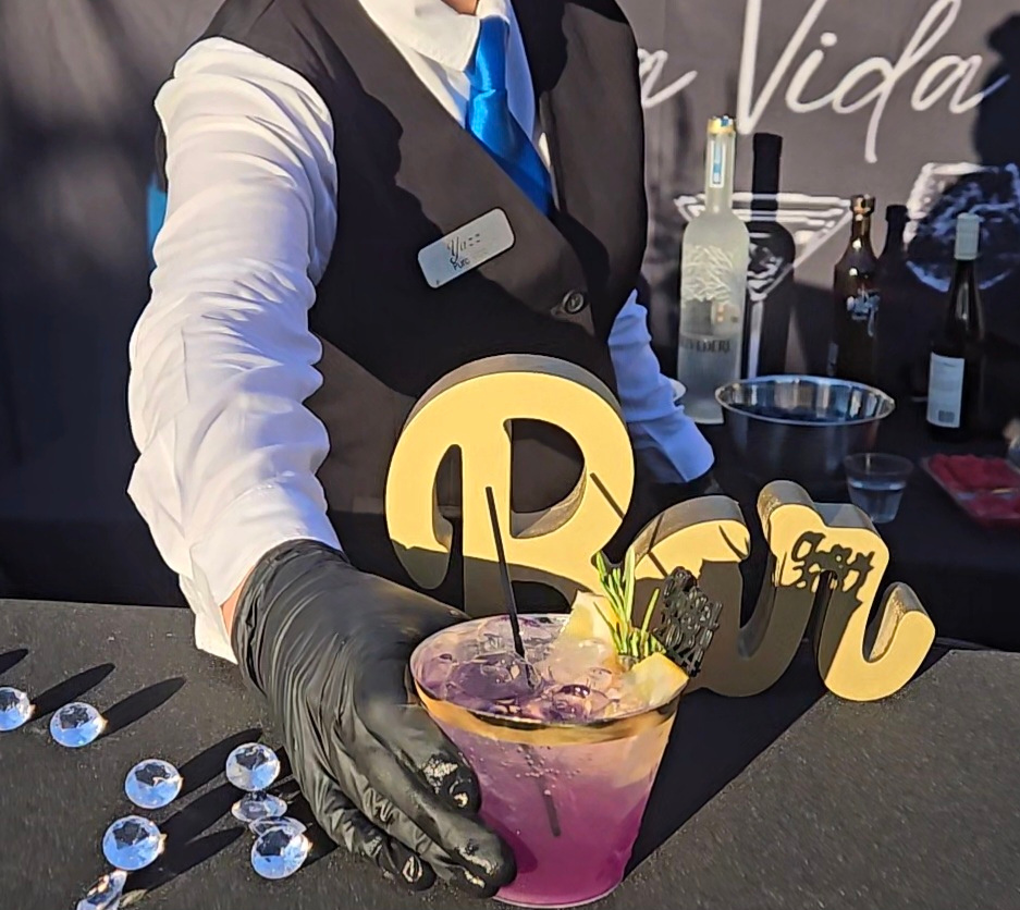 Person in formal attire serving a pink cocktail with decorative signs, black gloves, and bar items in background.