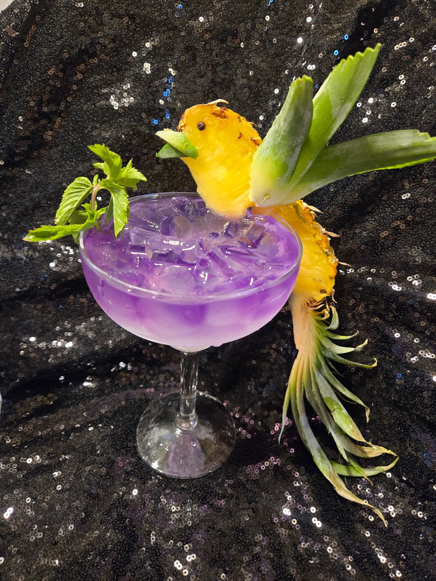 A colorful cocktail glass with purple liquid, decorated with a yellow bird, green leaves, and a pineapple tail, on a black background.
