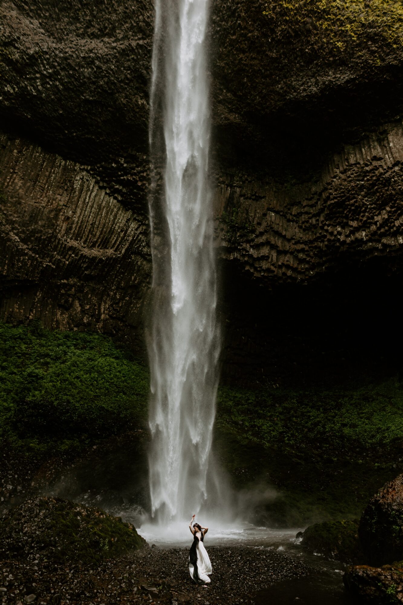 Person standing at the base of a tall waterfall inside a dark cave, surrounded by mossy rocks and greenery.