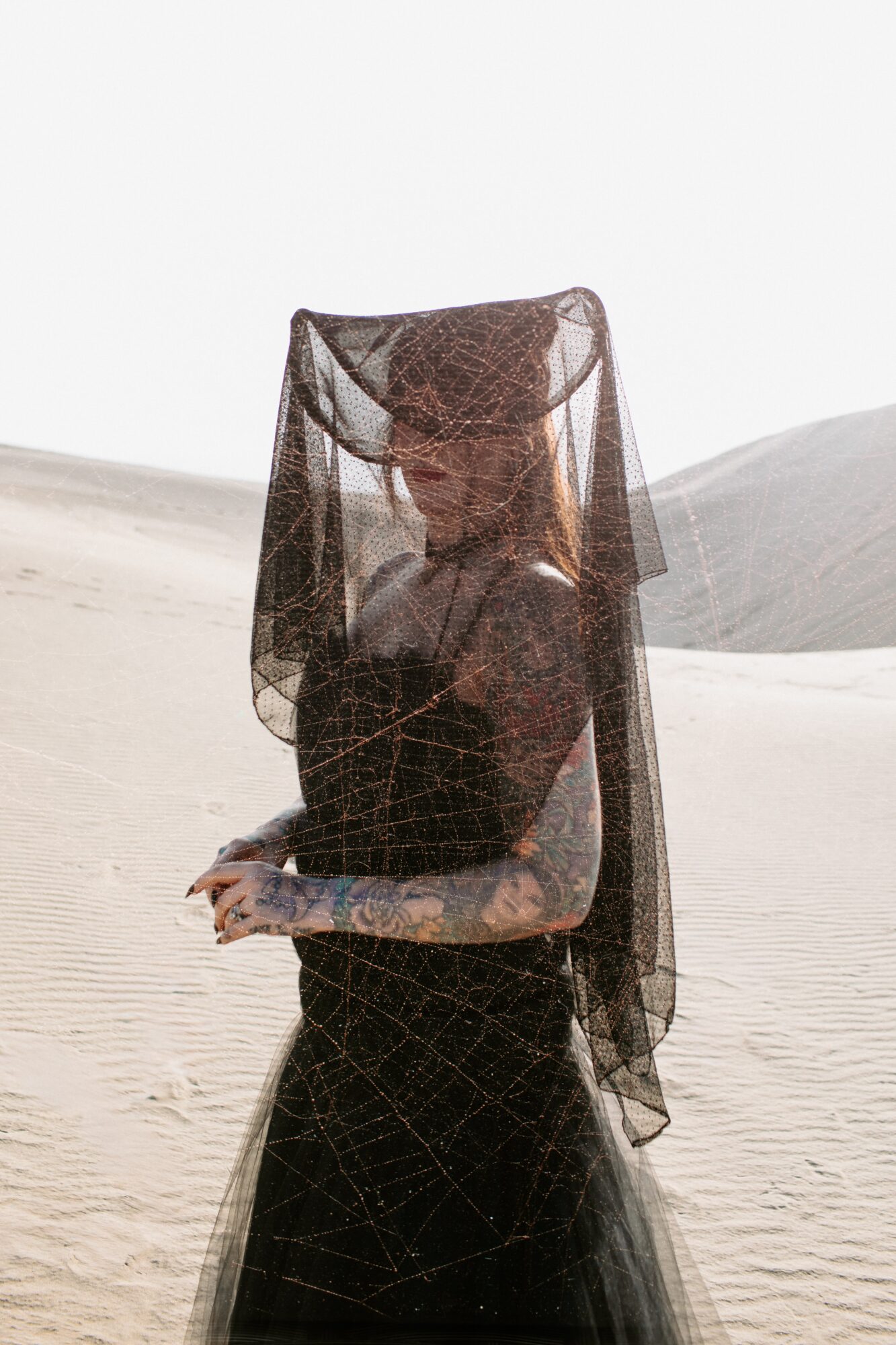 Person with tattoos wearing a black dress and veil in a desert landscape, facing sideways.