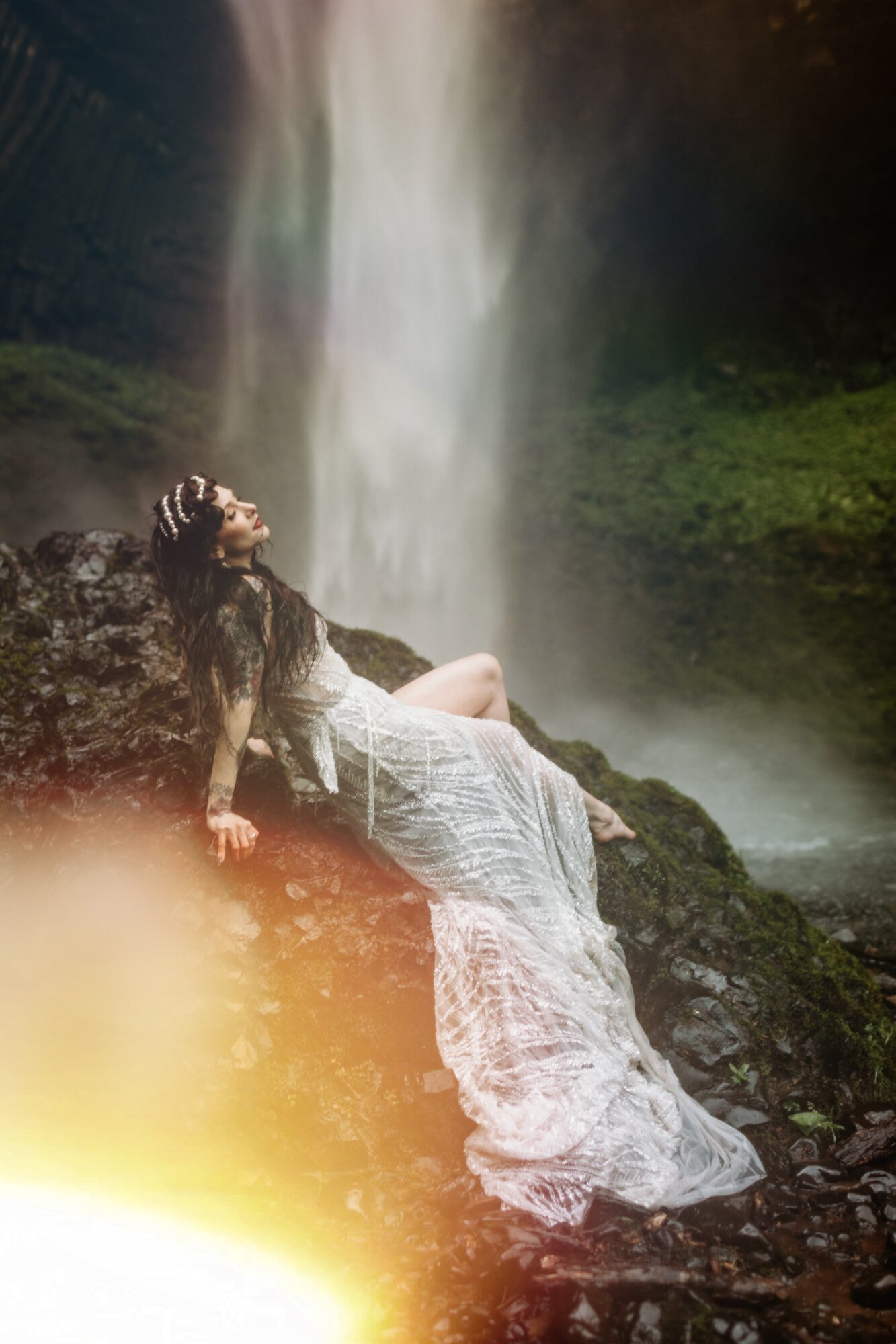 Woman in a white dress reclining on rocks near a waterfall with mist and greenery in the background.