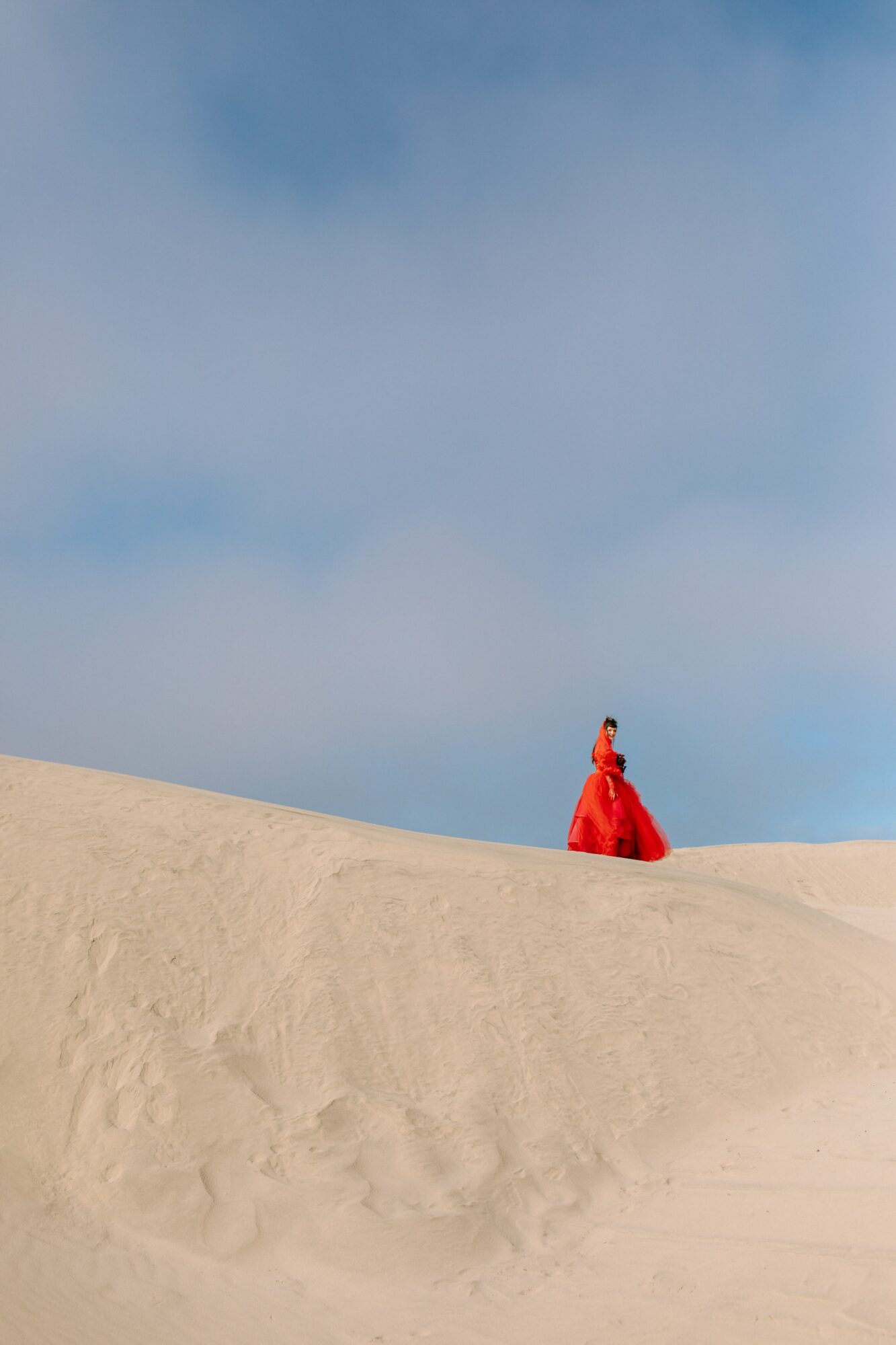 Person in a red dress walking on sand dunes under a blue sky with clouds.
