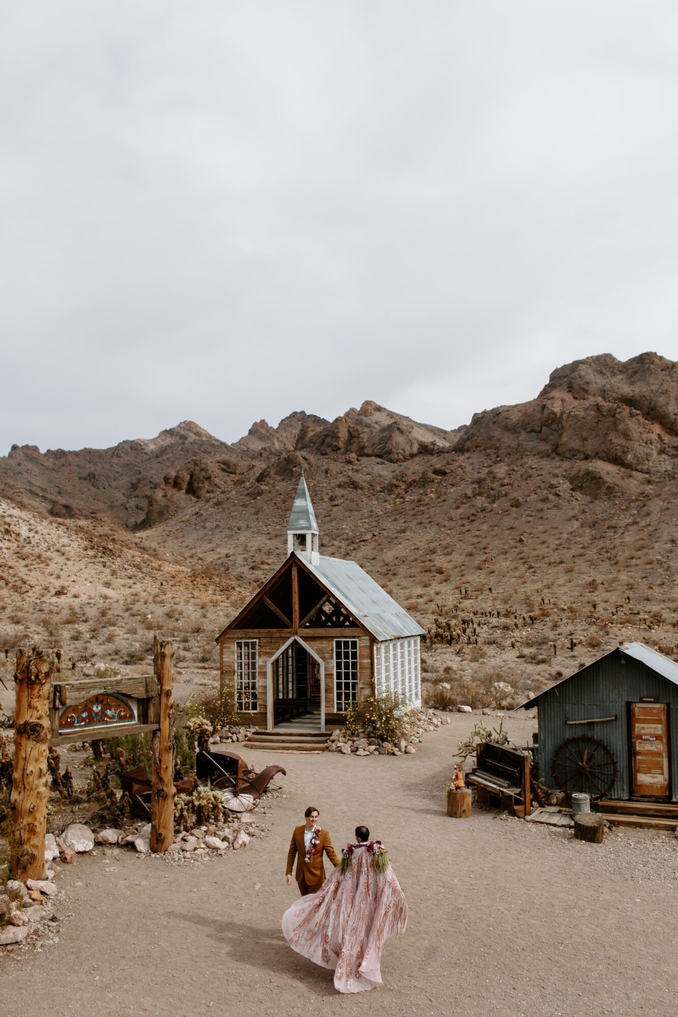 Two women in pink dresses walk toward a small church with a steeple, set against rocky mountains and cloudy sky.