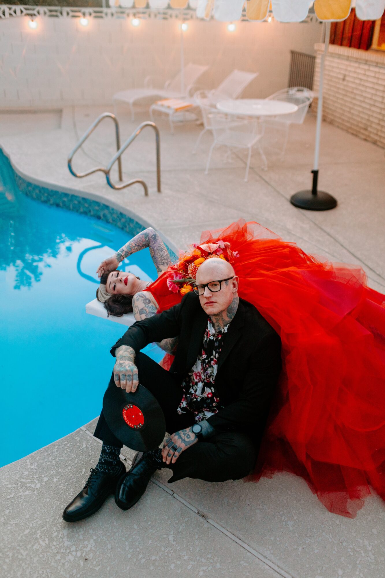 Person in a red gown and glasses sitting by a swimming pool with lounge chairs in background.