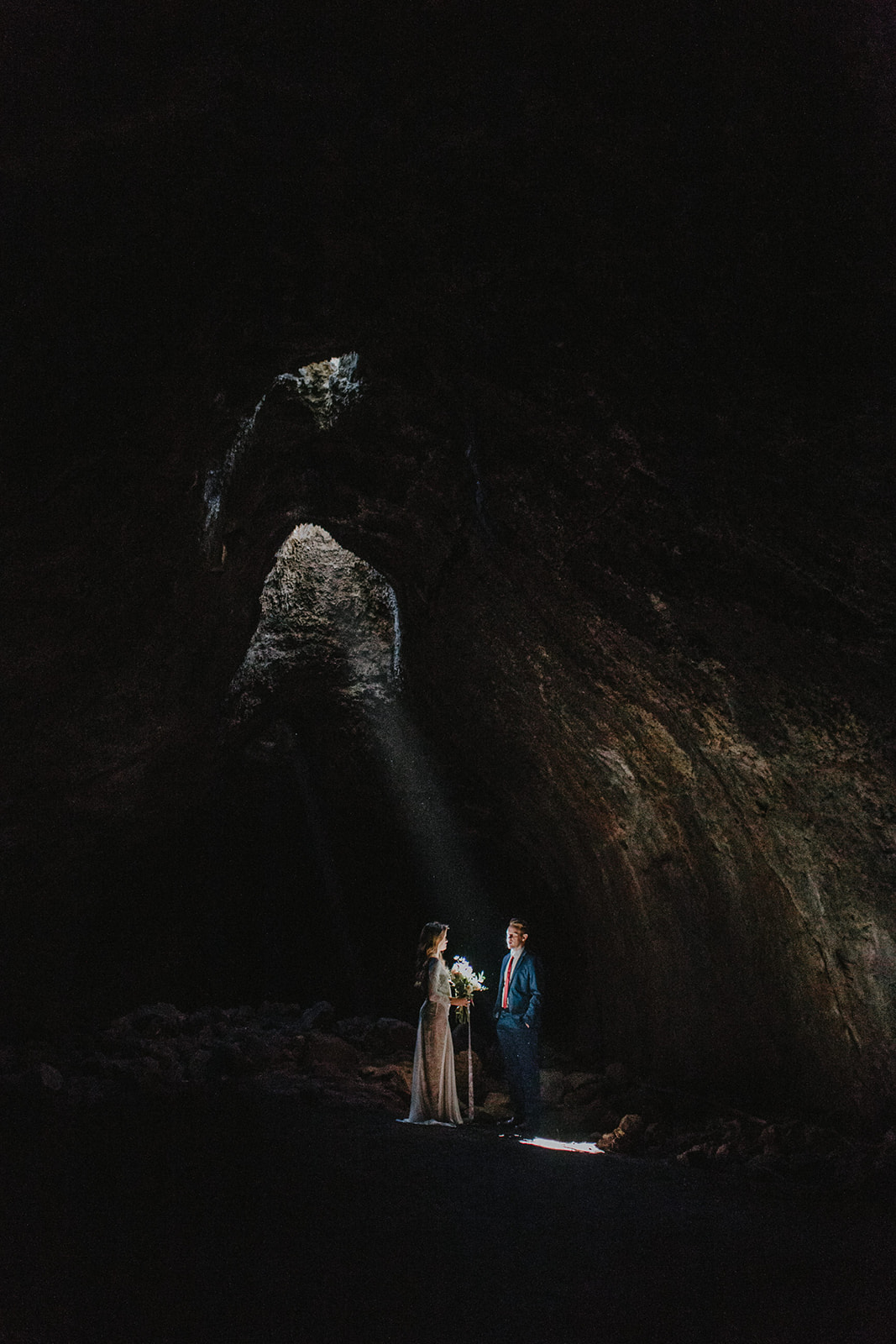 Two people stand inside a dark cave with a small opening letting light in, illuminating the scene.