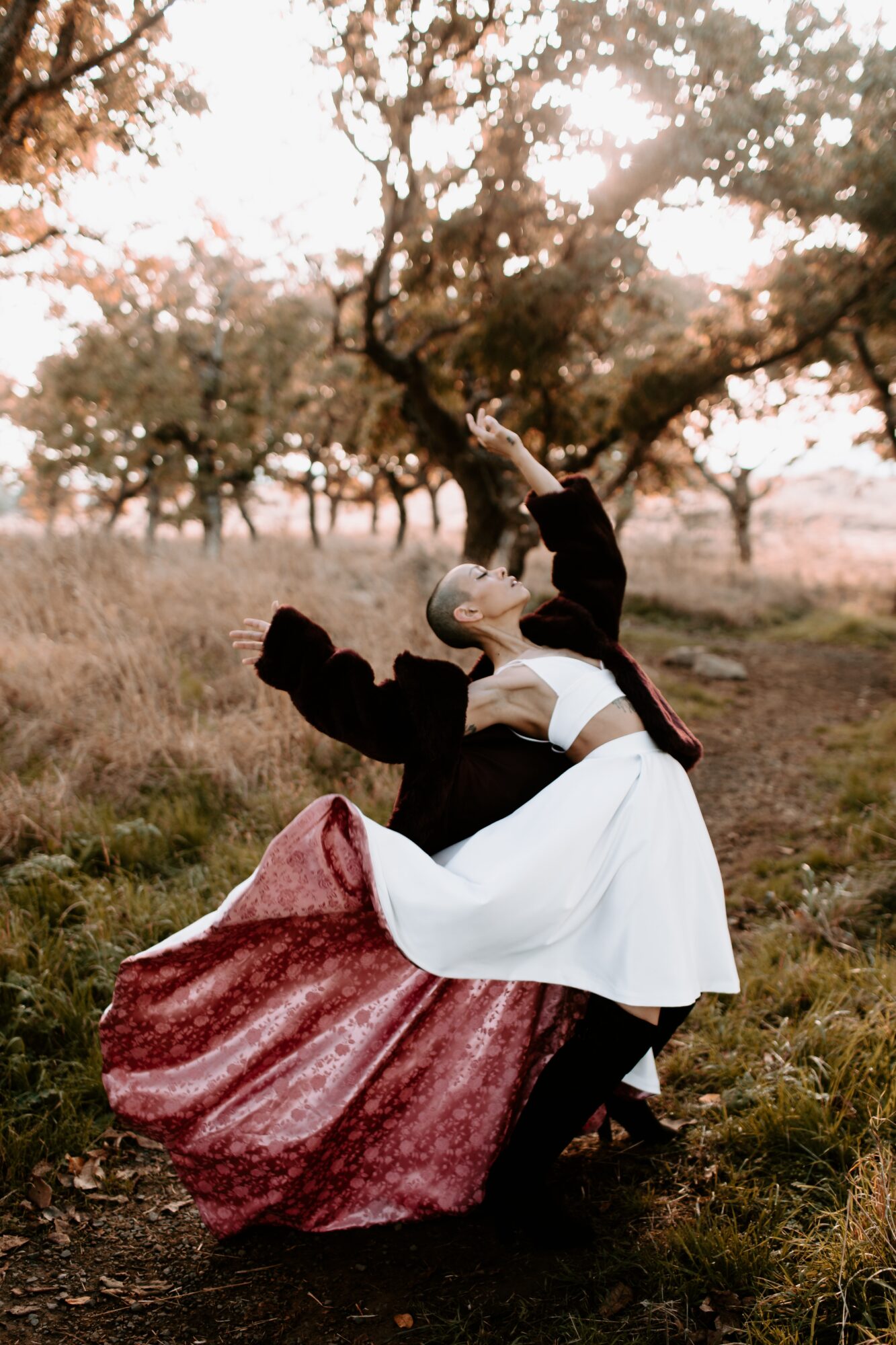 A couple dancing outdoors in a field with trees, the woman wearing a white dress and the man in dark clothing.