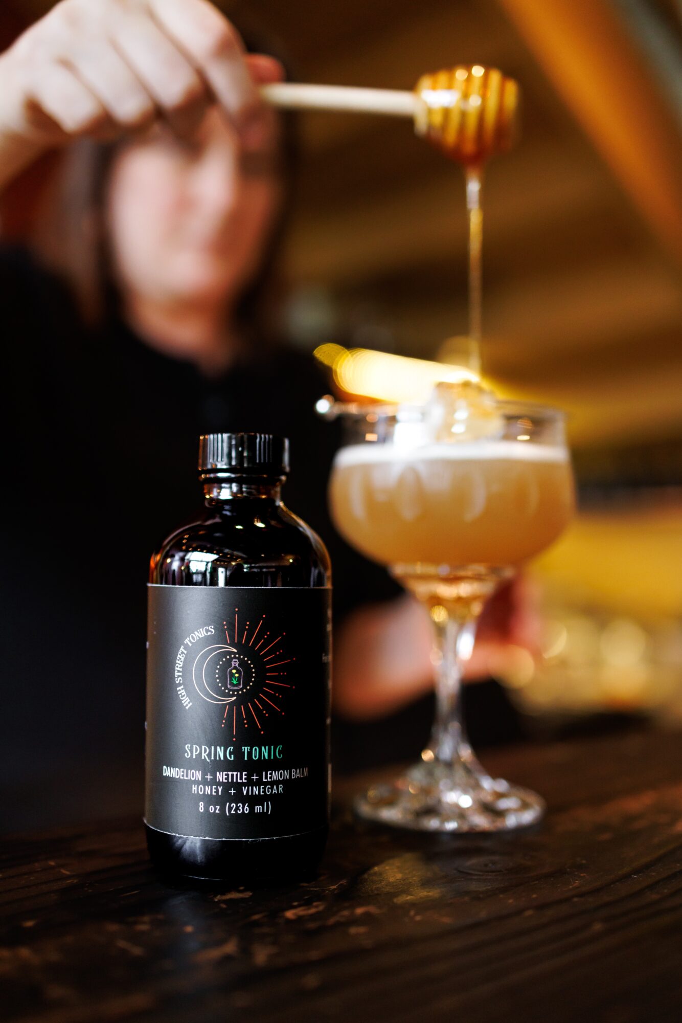 Person drizzling honey into a cocktail glass with a bottle of spring tonic nearby, blurred background.