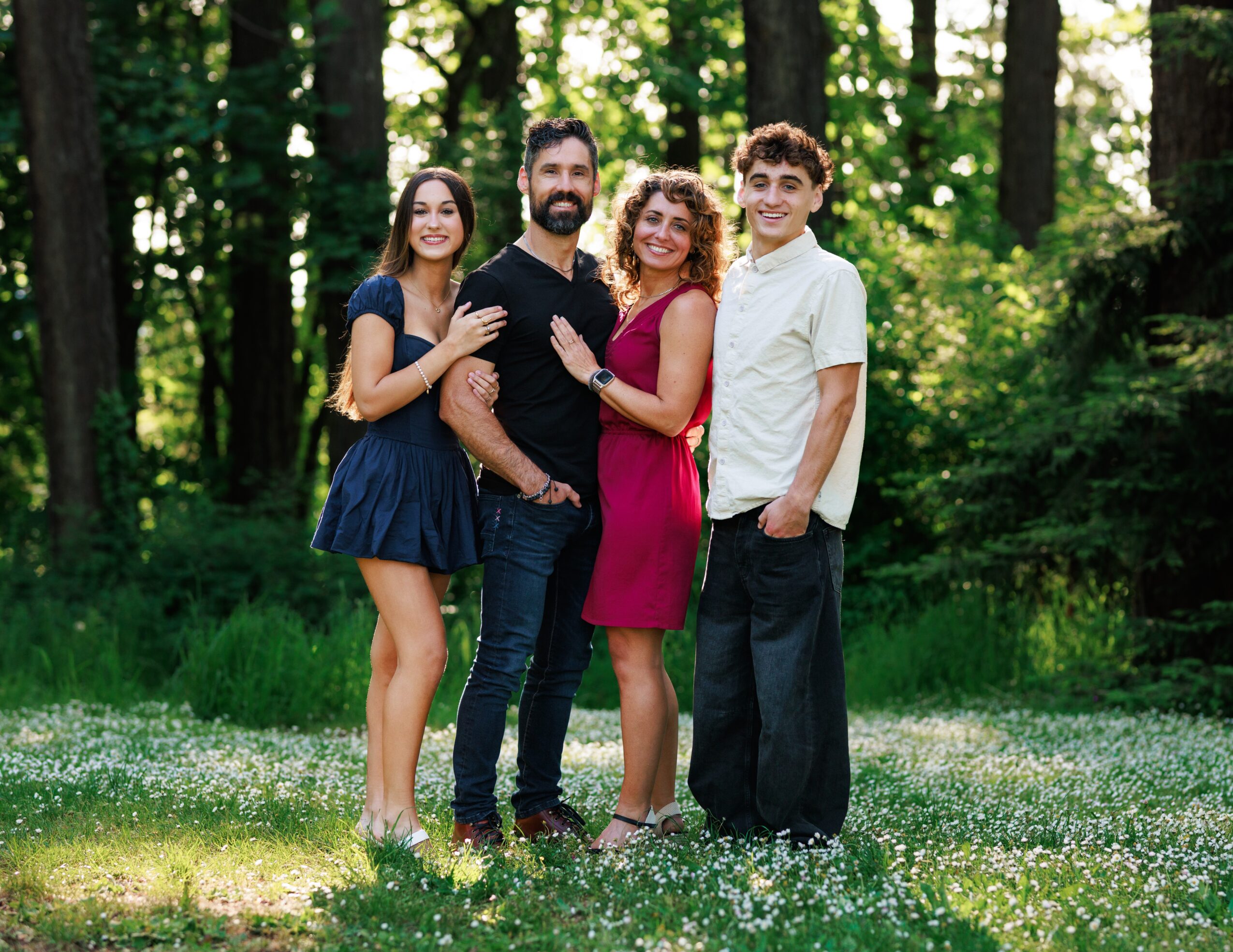 Four people standing outdoors in a forest, smiling, with trees and grass in the background.