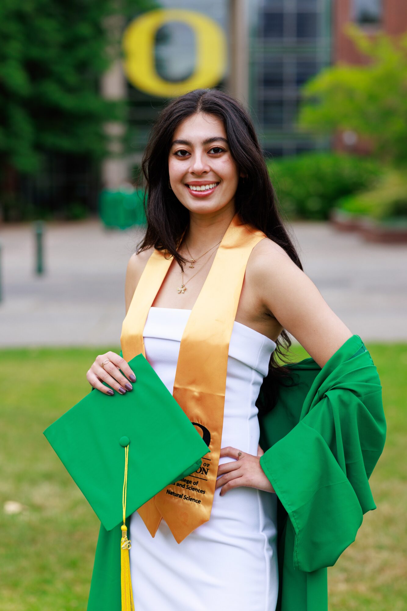 Young woman in graduation gown and sash smiling outdoors, holding a green cap, with trees and buildings in background.