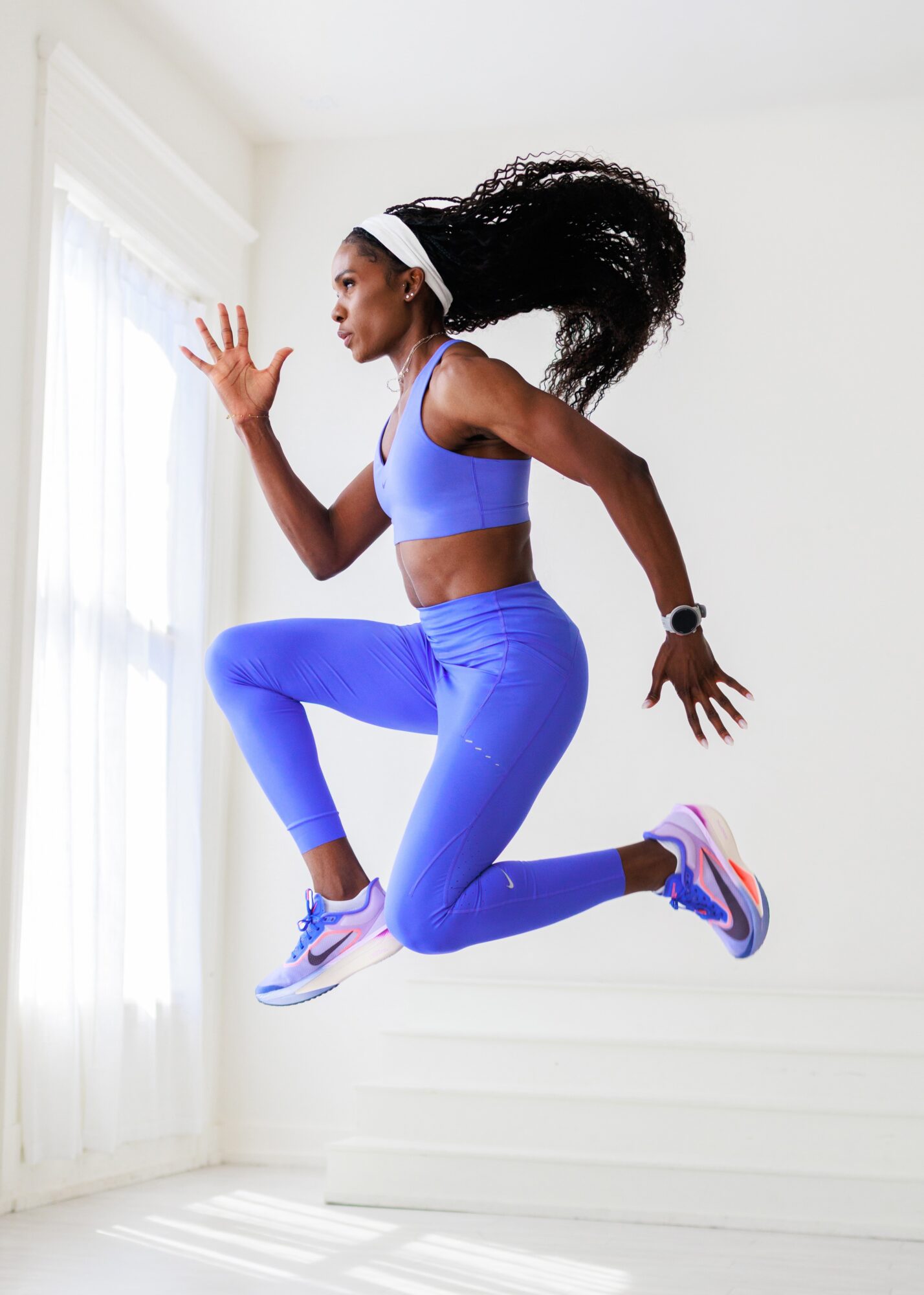 Woman in blue athletic wear jumping indoors near a window, with her hair flowing and arms raised.