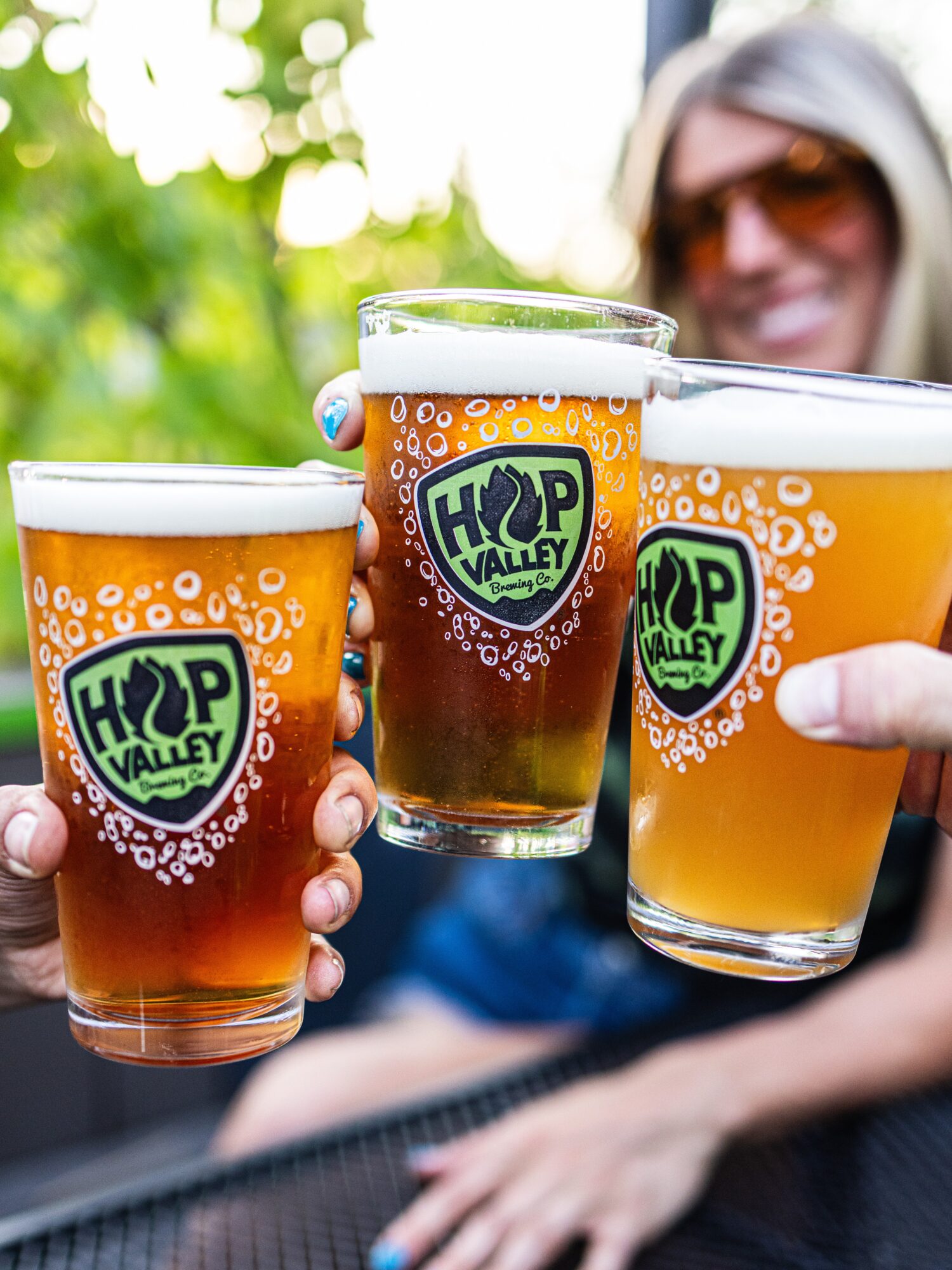 Three people holding glasses of beer outdoors with green foliage in background, smiling, wearing sunglasses.