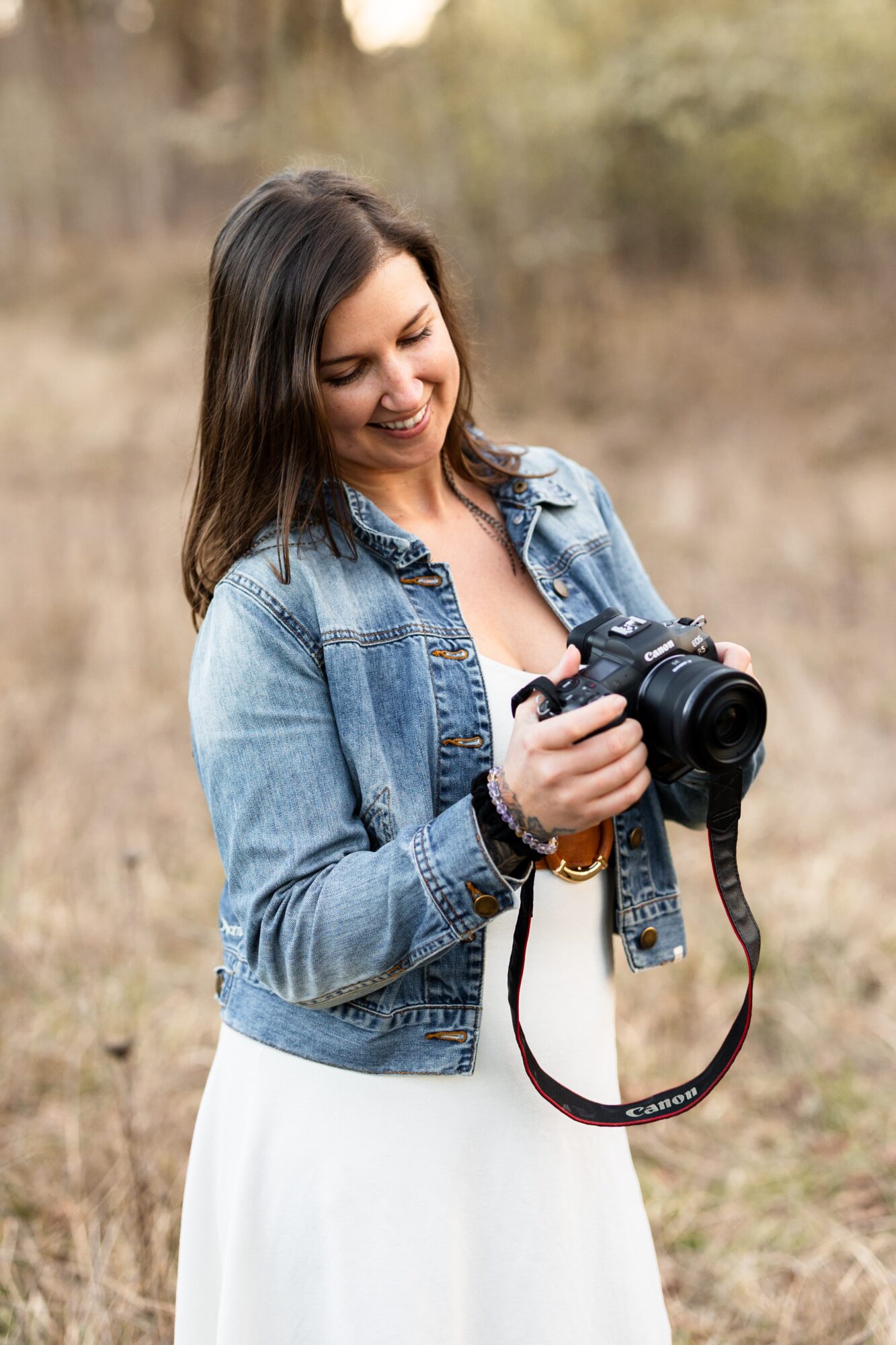 Young woman in denim jacket holding a camera outdoors, smiling, with blurred natural background.