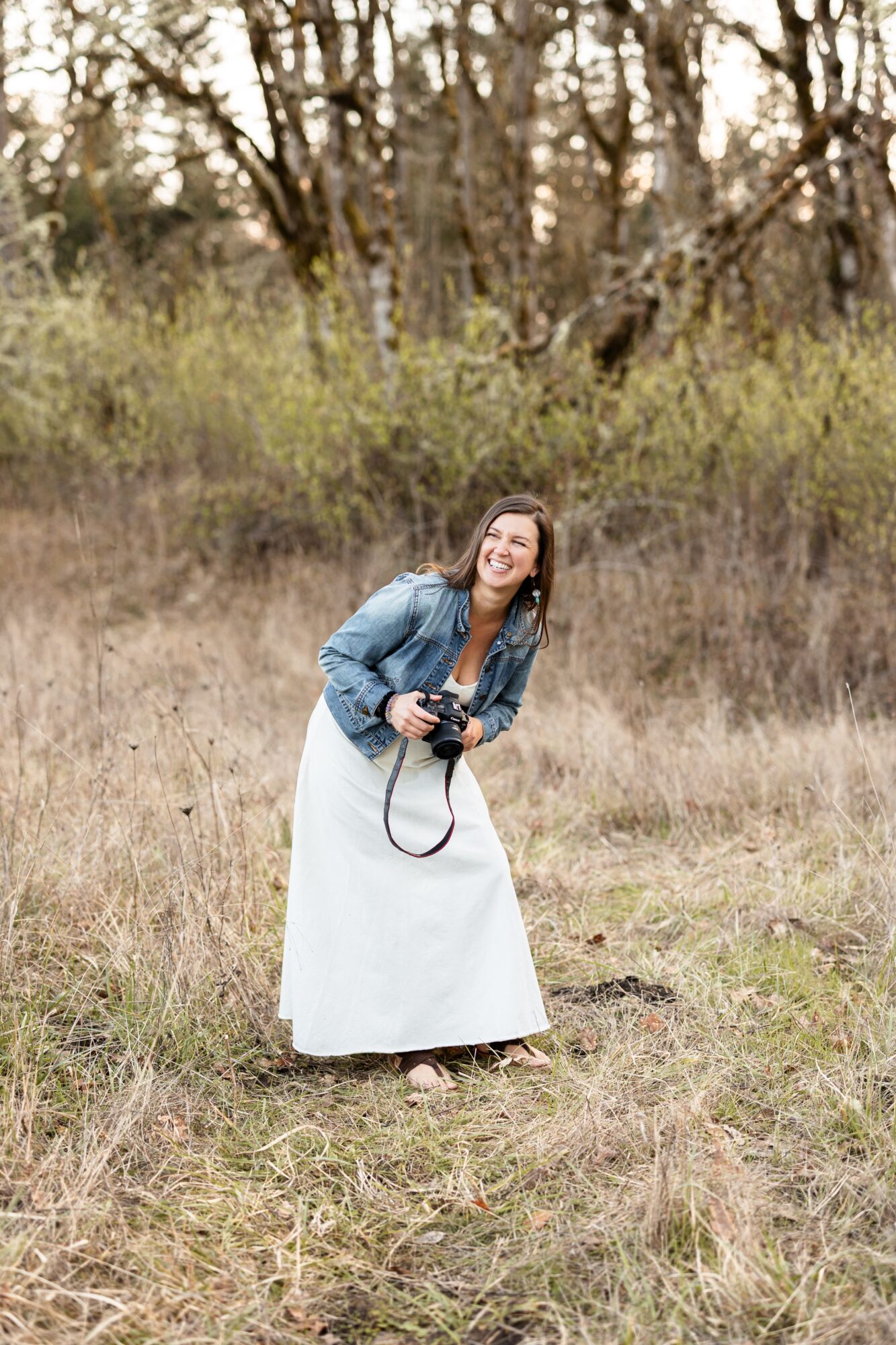Woman standing outdoors in a grassy field, smiling, holding a camera, with trees in the background.