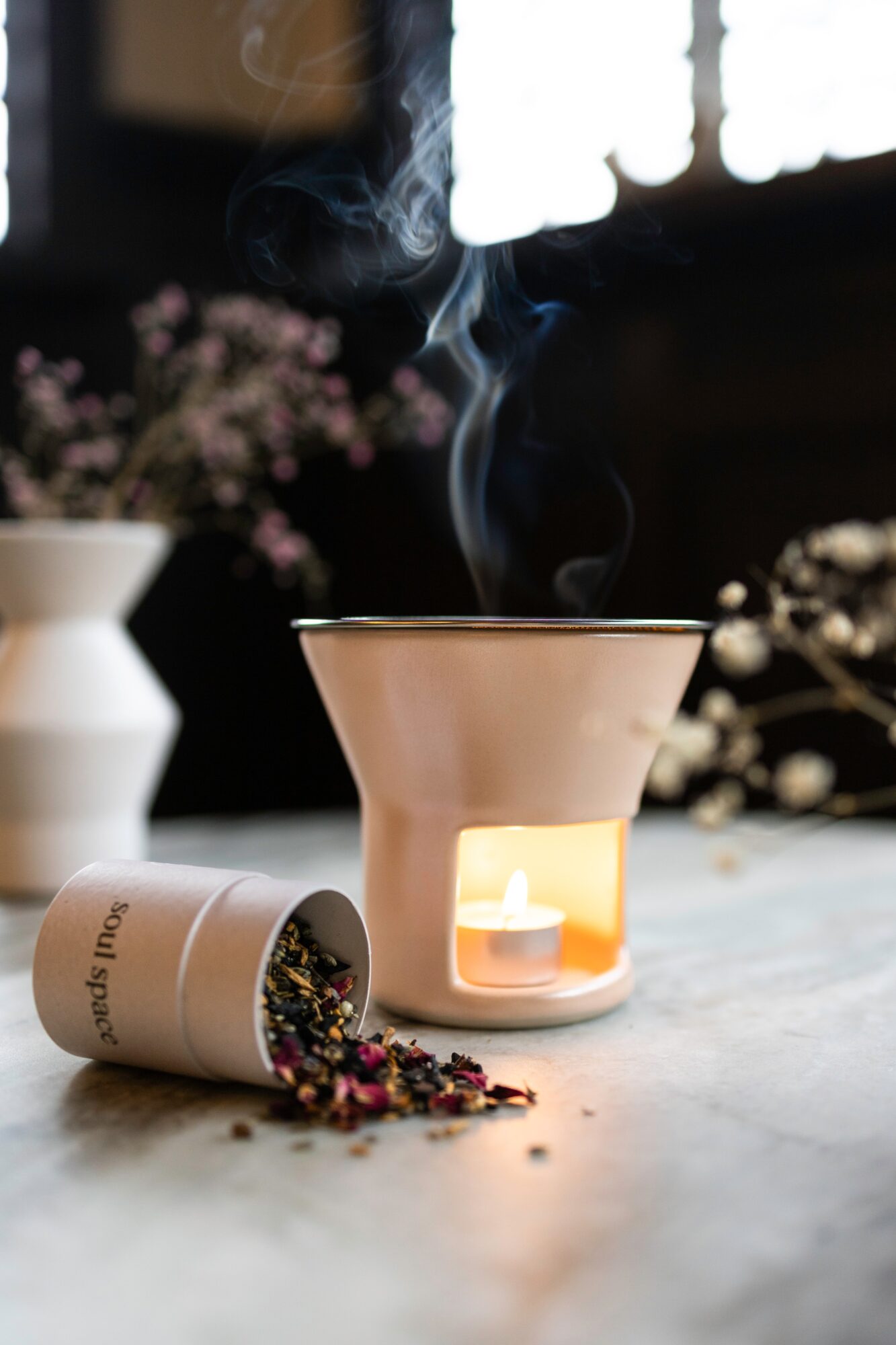 A ceramic essential oil diffuser with a small candle inside, emitting steam, on a table with scattered herbs and flowers.
