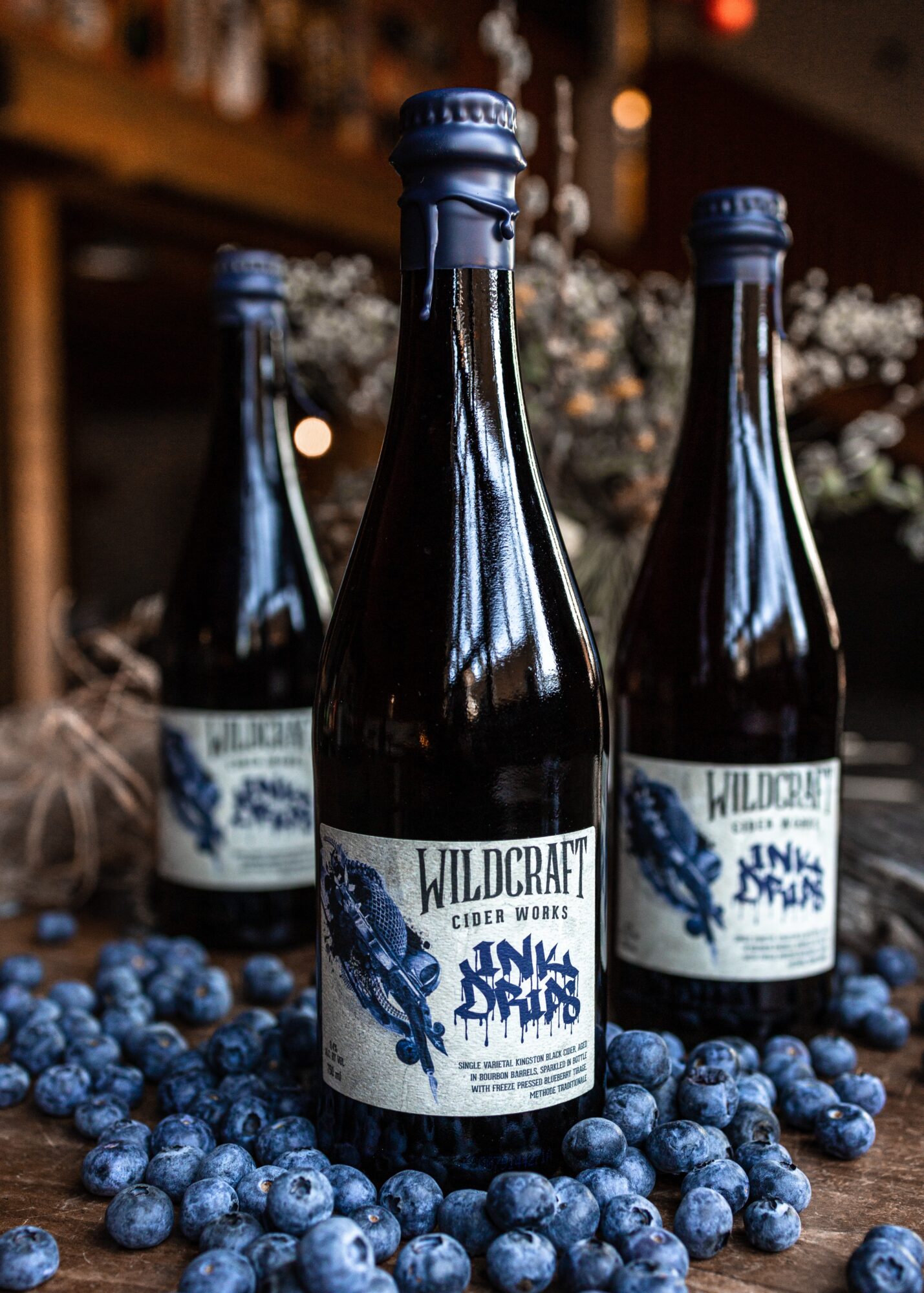Three bottles of Wildcraft cider surrounded by blueberries, with a blurred background of a cozy indoor setting.