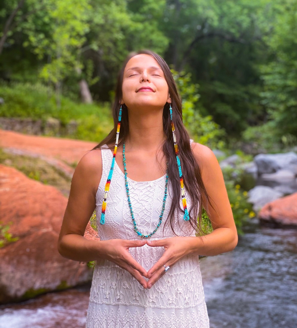 Young woman with long hair and colorful beads in a white dress, standing outdoors with eyes closed, smiling, near a stream.